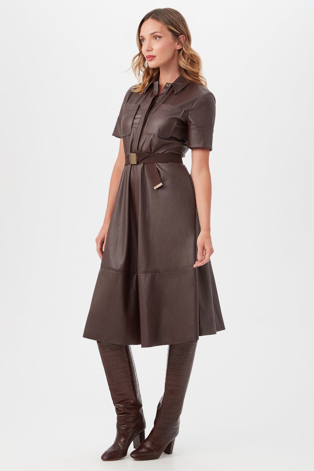 A woman stands against a plain white background wearing the Misola DressÃ¢â‚¬â€a dark brown, short-sleeve, collared leather dress with a belted waistÃ¢â‚¬â€paired with matching knee-high heeled boots.