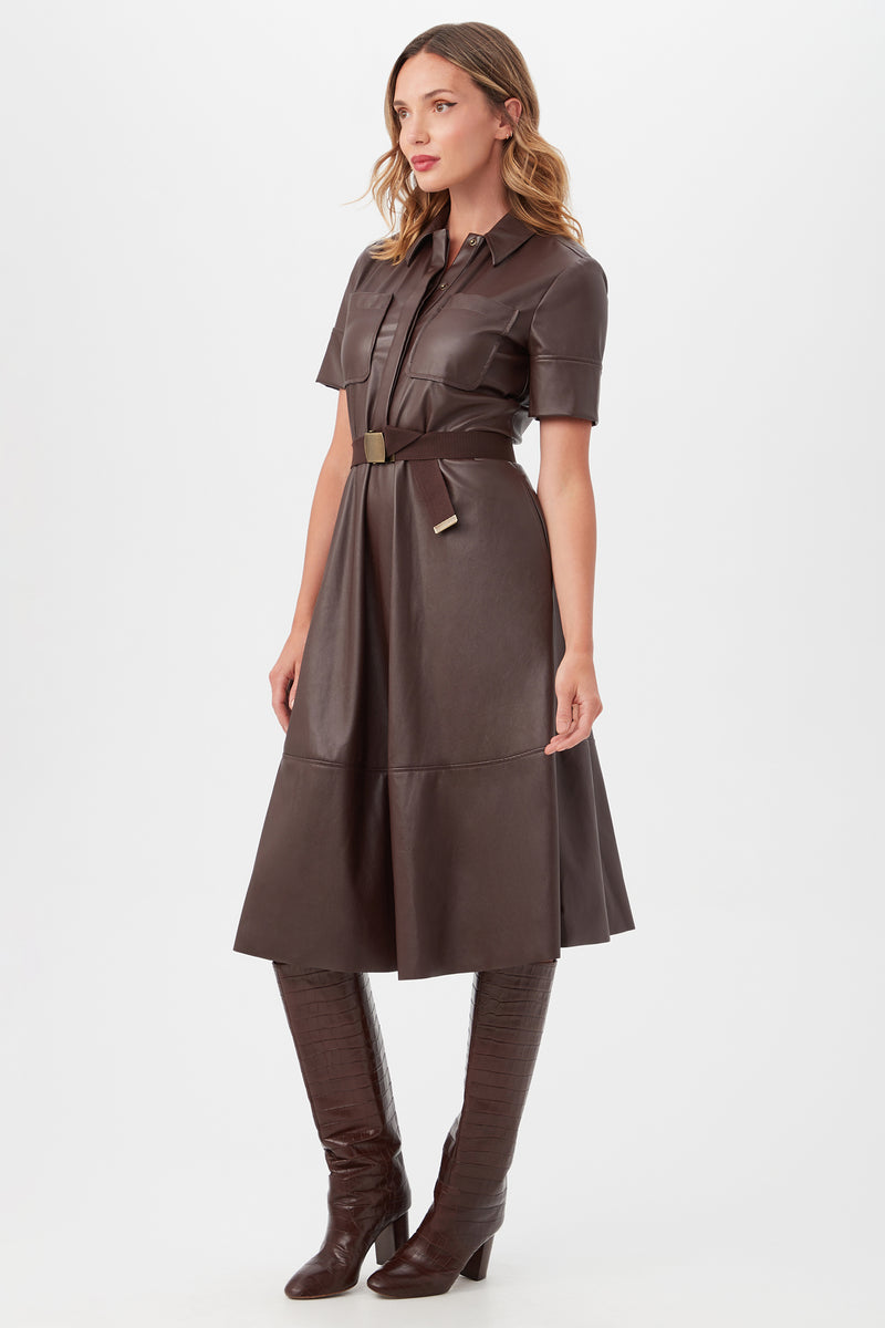A woman stands against a plain white background wearing the Misola DressÃ¢â‚¬â€a dark brown, short-sleeve, collared leather dress with a belted waistÃ¢â‚¬â€paired with matching knee-high heeled boots.