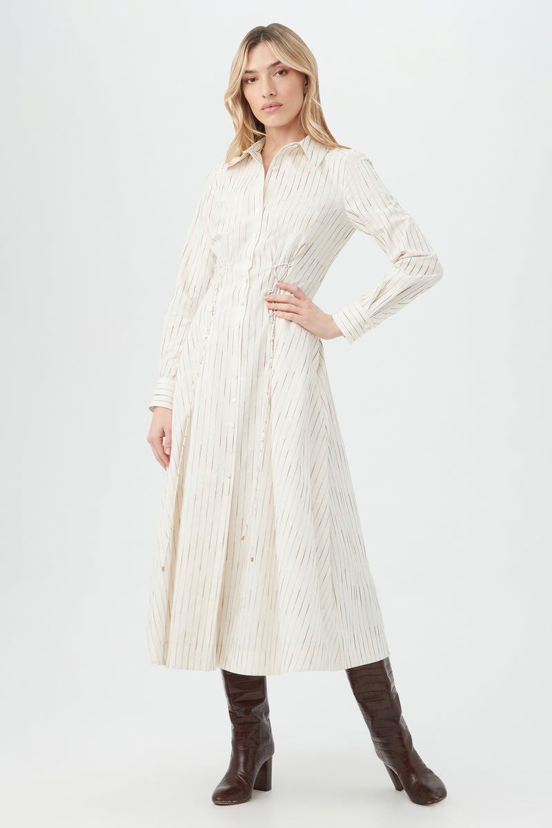 A woman models the Omari Dressâ€”an ivory, ankle-length long-sleeve dress with subtle vertical stripesâ€”paired with dark brown knee-high boots, posing against a plain background.