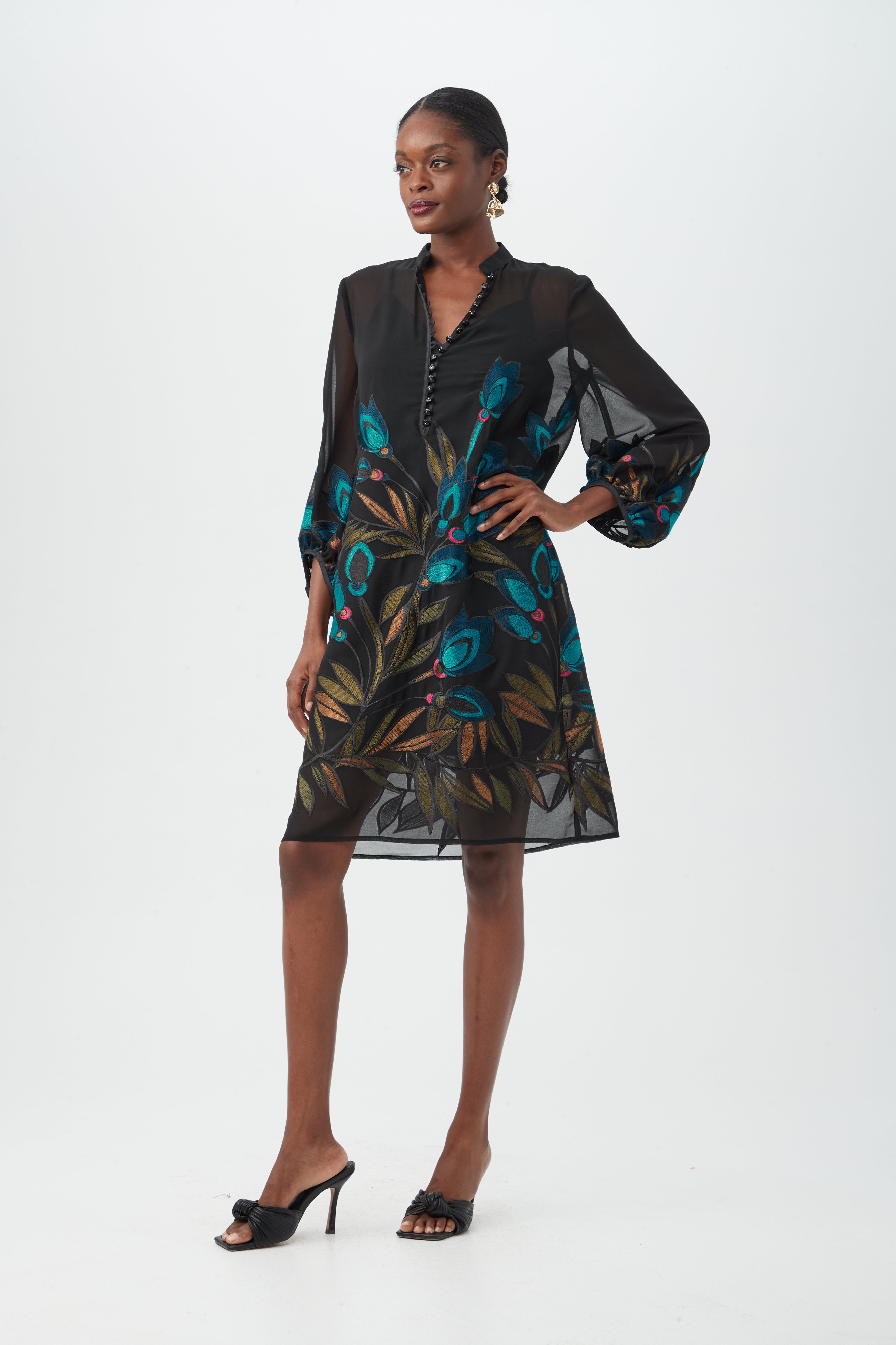 A woman poses confidently against a white background, wearing the Boheme 2 Dressâ€”a black sheer dress with colorful floral patterns and puffed long sleevesâ€”paired with black heeled sandals and her hair pulled back.