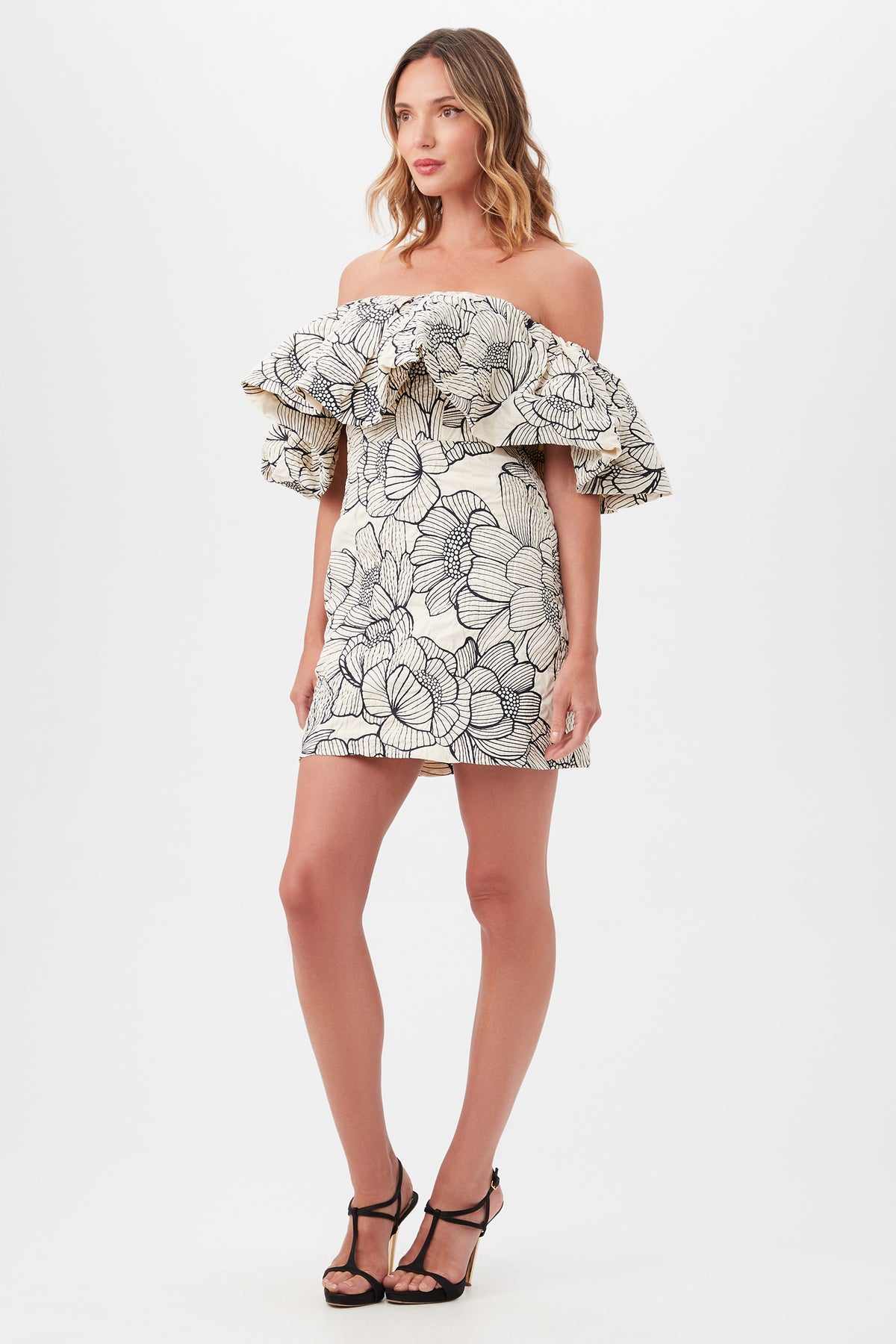 Woman wears the Nia Dressâ€”an off-the-shoulder white mini with bold black floral print and a ruffled necklineâ€”paired with black strappy heels, standing against a plain white background.