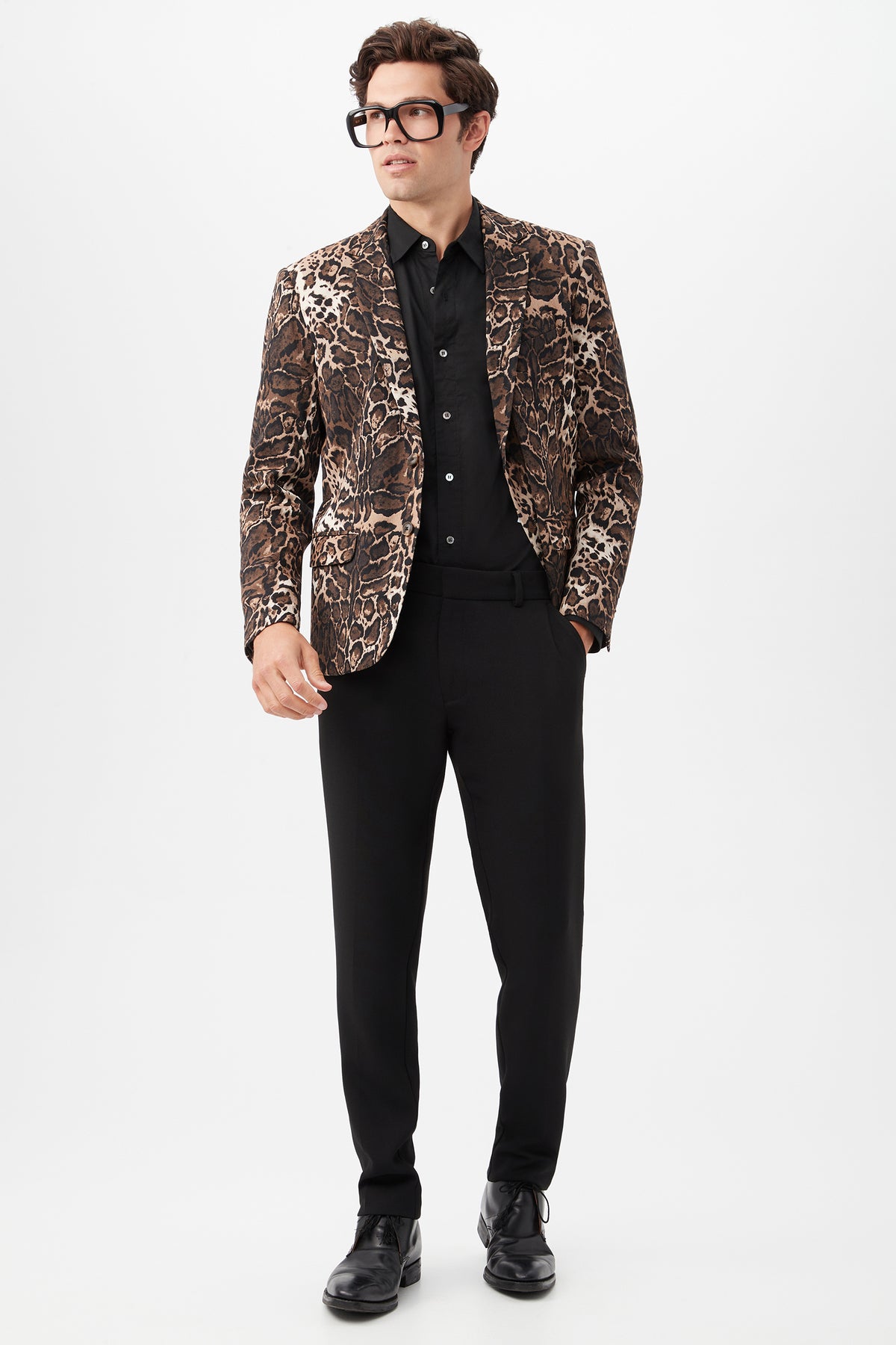 A man stands against a plain white background wearing black glasses, a black shirt, black shoes, a brown leopard-print blazer, and Clyde Slim Trouser - Hi-Line Suiting.