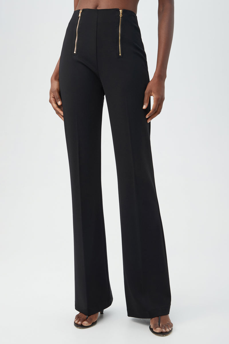 A person wears the Kiara Pant in blackâ€”high-waisted, wide-leg style with two gold front zippersâ€”paired with open-toe heels against a plain light background.