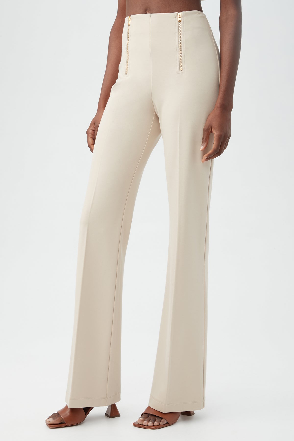 A person wears the Kiara Pantâ€”high-waisted, beige, wide-leg pants with two gold hip zippersâ€”paired with brown heeled sandals, standing against a plain white background.