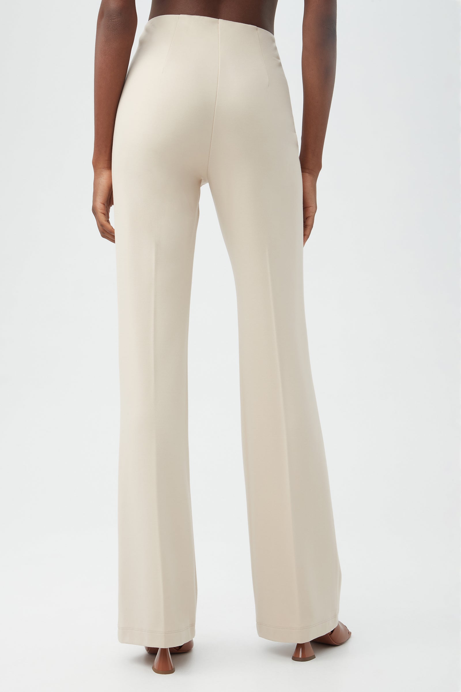 A person stands facing away, wearing the Kiara Pantâ€”high-waisted, light beige, wide-leg trousersâ€”and open-toe heels against a plain white background.