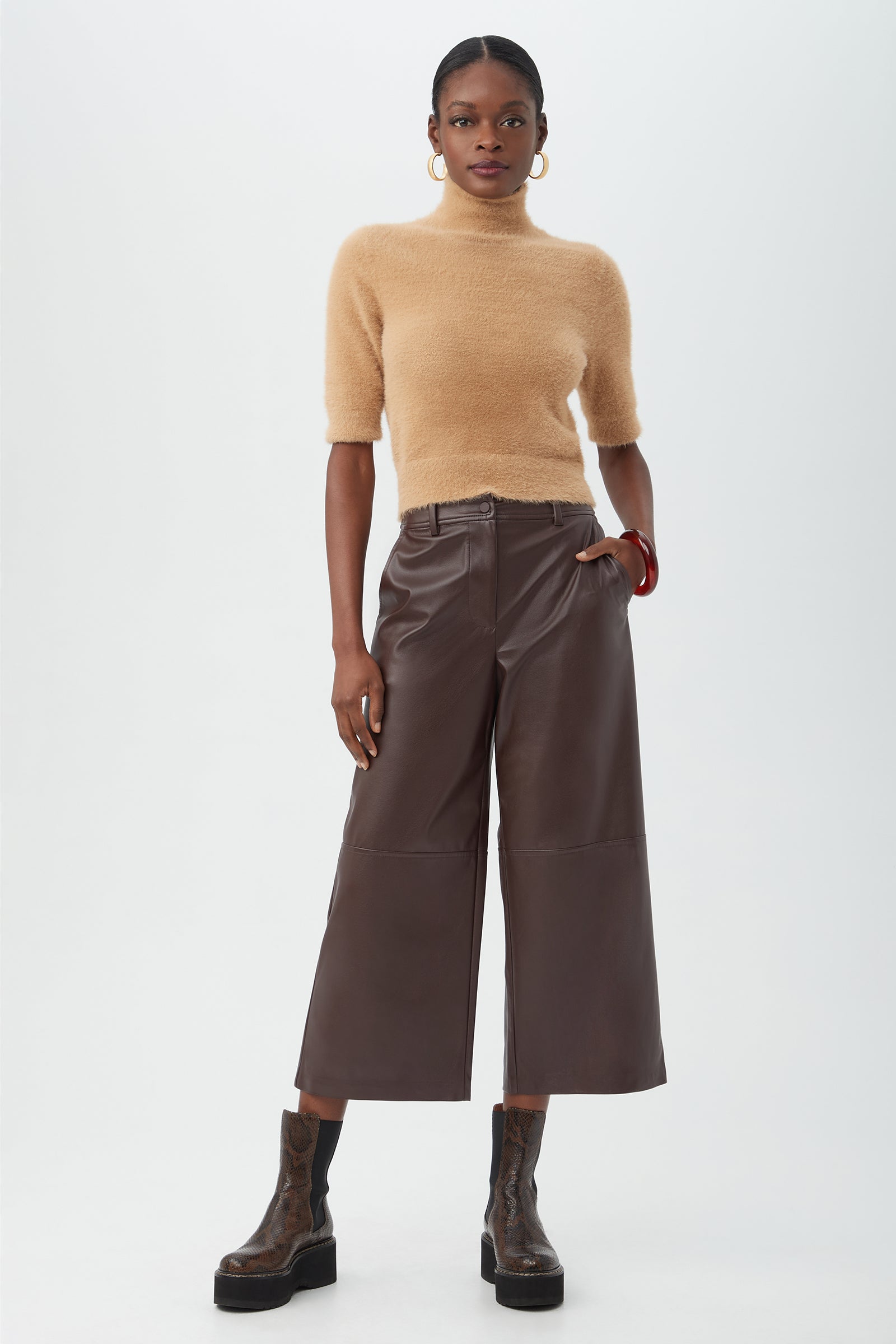 A woman stands against a white background wearing the Mali Fuzzy Sweater in camel, paired with wide-leg brown leather cropped pants, brown platform boots, large hoop earrings, and a red bangle.