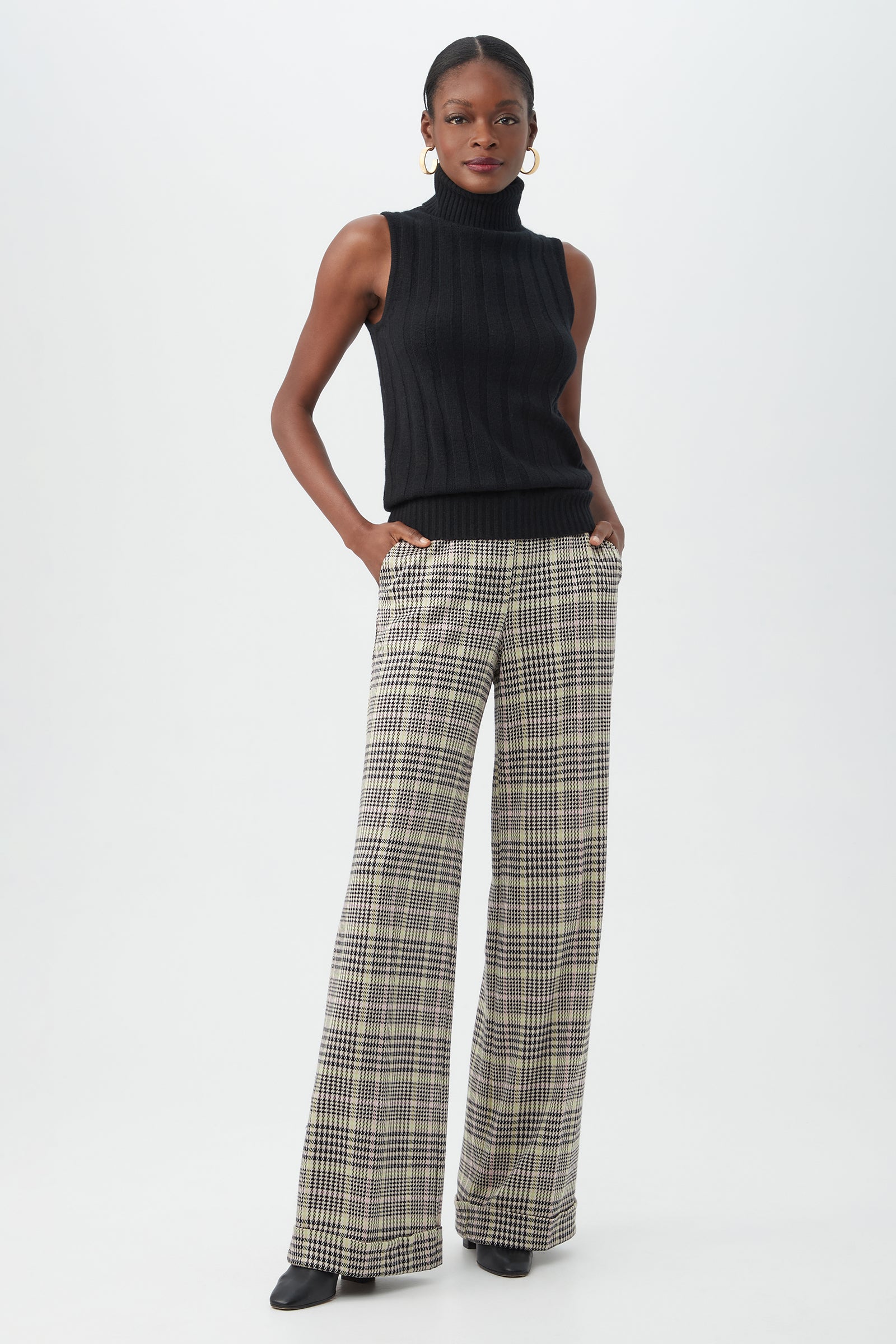 A woman stands against a plain background wearing the Onassis Knit Top, high-waisted plaid wide-leg pants, black pointed shoes, gold hoop earrings, and has her hands in her pockets.