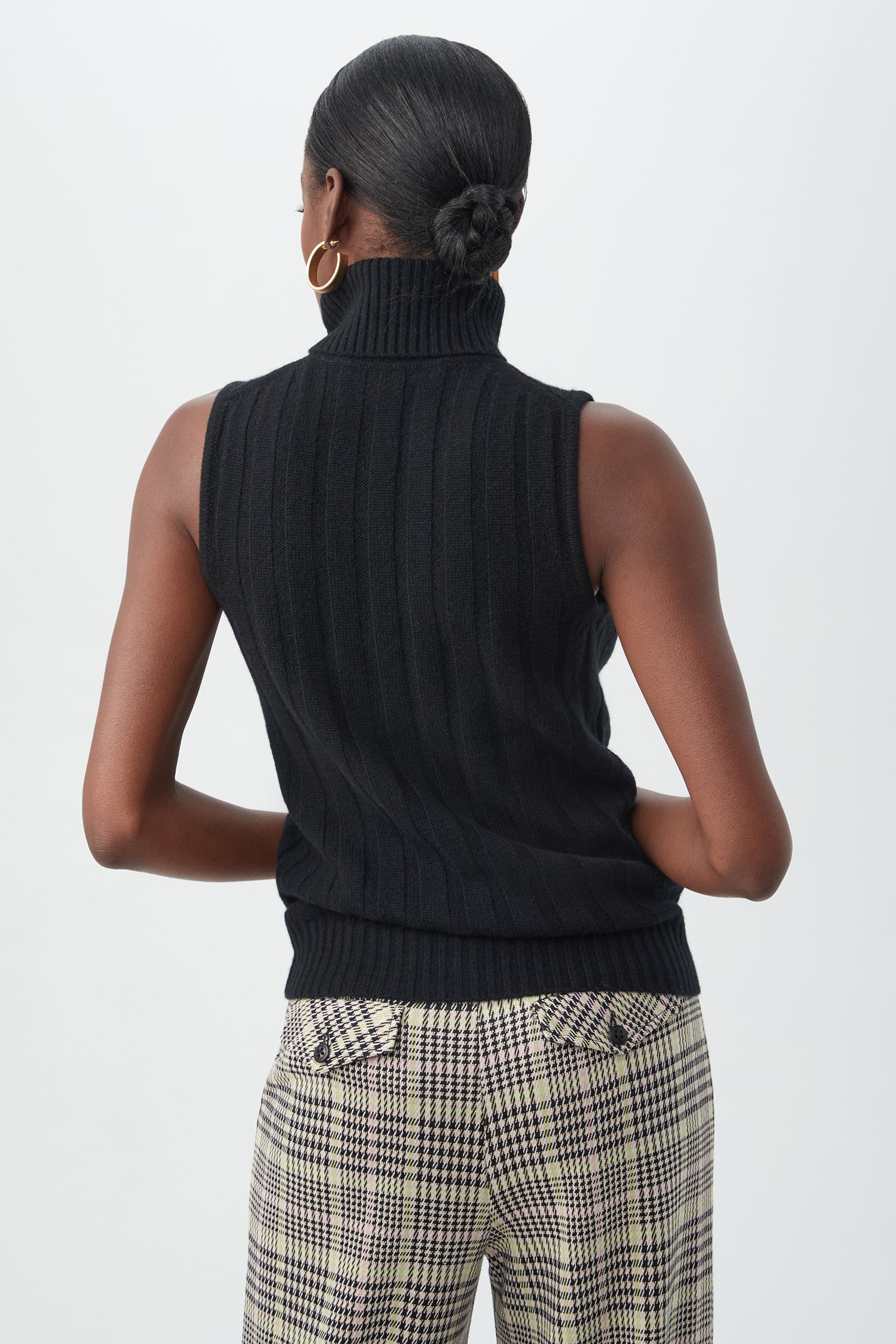 A woman with her hair in a low bun is shown from the back against a plain light background wearing the sleeveless black Onassis Knit Top and plaid pants.