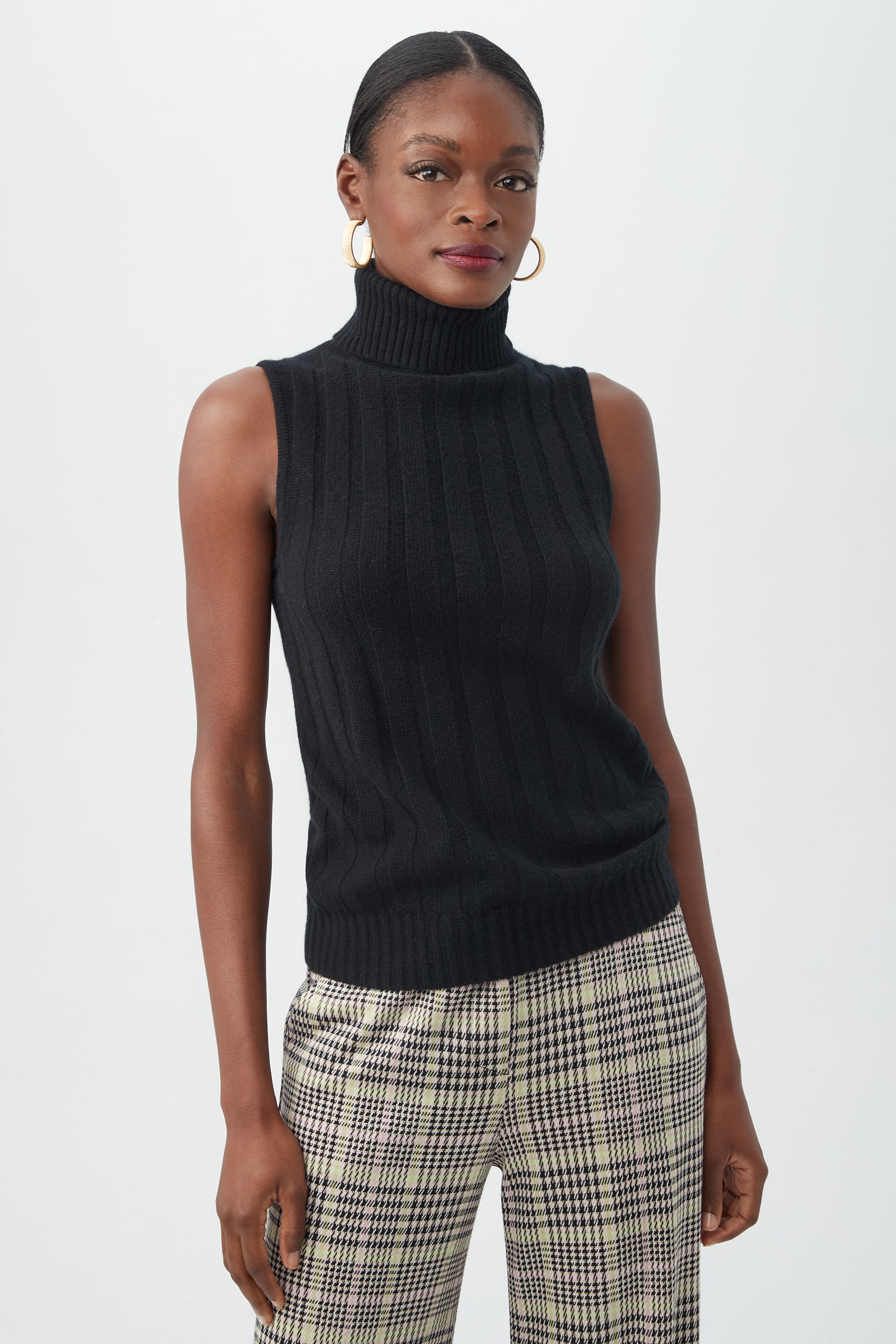 A woman stands against a plain light background wearing the black Onassis Knit Top, paired with plaid trousers and gold hoop earrings. Her hair is pulled back, and she has a confident expression.