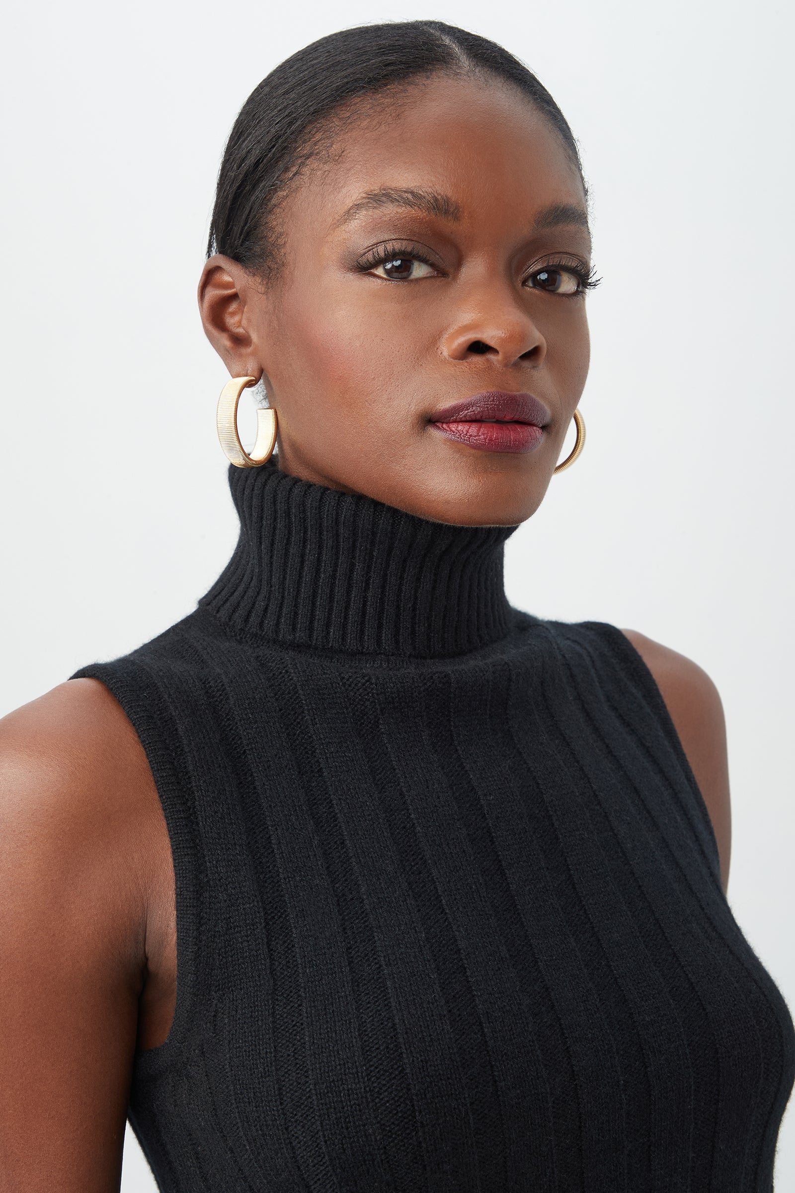 A woman with dark skin and pulled-back hair models the sleeveless black Onassis Knit Top, featuring a ribbed turtleneck, gold hoop earrings, and a confident gaze against a plain light background.