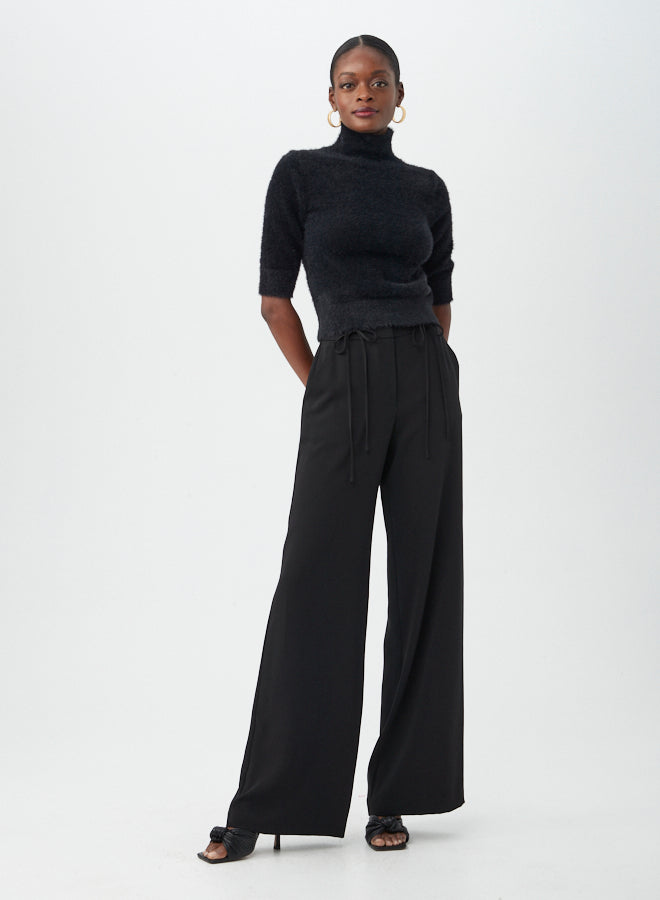 A woman stands against a white background wearing the Mali Fuzzy Sweater, black wide-leg tie-waist pants, black heeled sandals, and gold hoop earrings.