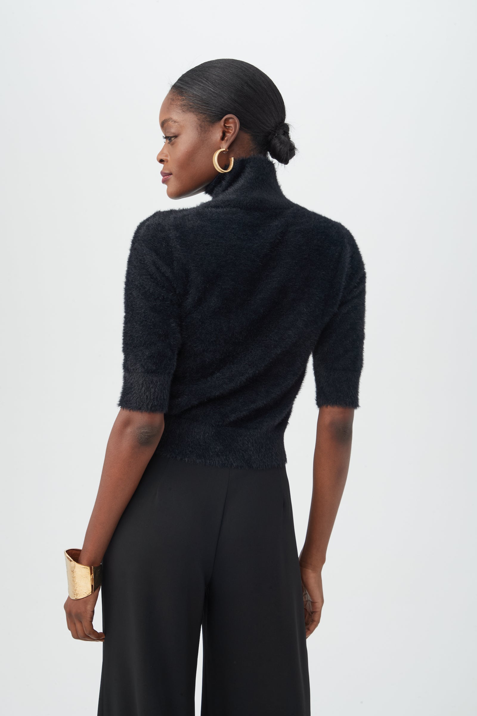 A woman, hair in a neat bun, faces away wearing the Mali Fuzzy Sweater—a black short-sleeve turtleneck—paired with black wide-leg pants, large gold hoop earrings, and a gold cuff bracelet against a plain background.
