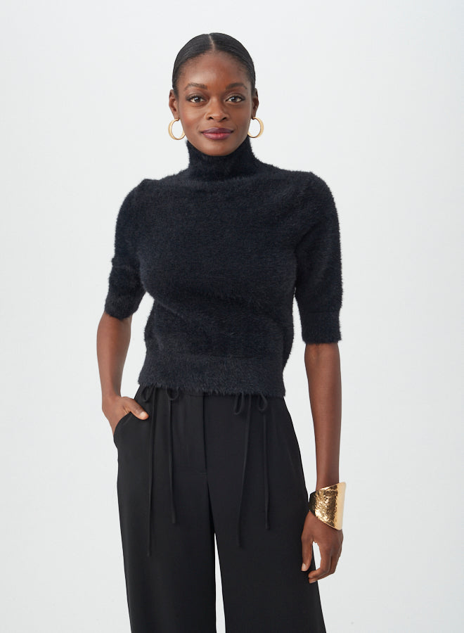 A woman smiles slightly against a plain background, wearing the black Mali Fuzzy Sweater with short sleeves and a turtleneck, paired with wide-leg black pants, gold hoop earrings, and a large gold cuff bracelet. One hand is in her pocket.