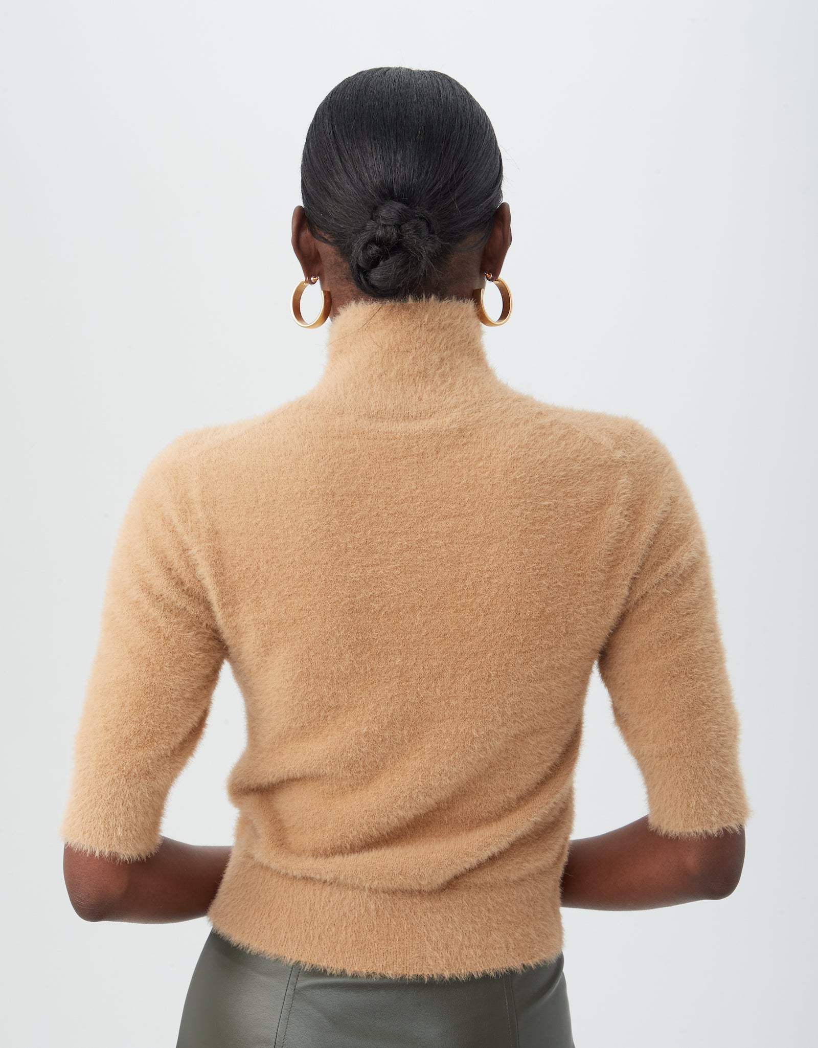 A person with neatly styled hair in a low bun wears the Mali Fuzzy Sweater, a light brown turtleneck with half sleeves, paired with gold hoop earrings, standing against a plain light background facing away from the camera.