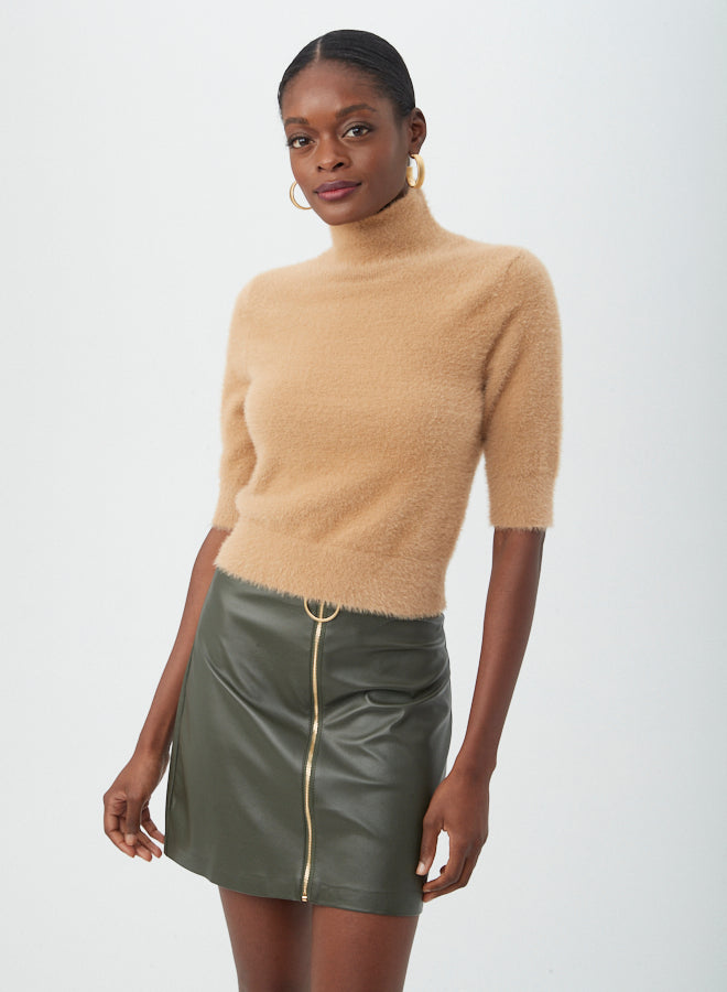 A woman wears the Mali Fuzzy Sweater, a tan turtleneck with short sleeves, paired with an olive green leather mini skirt with a front zipper, gold hoop earrings, and her hair pulled back against a light background.