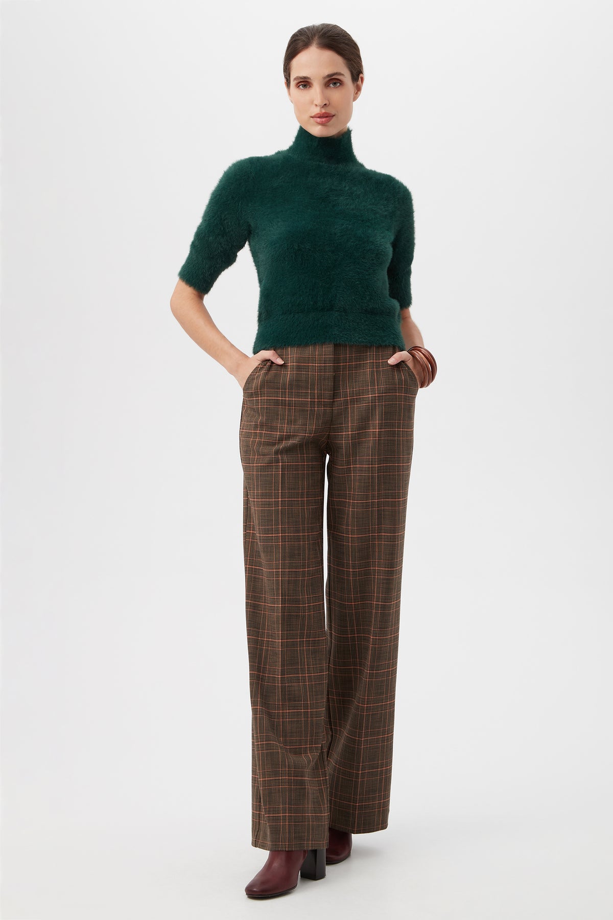 A woman stands against a white background, hands in pockets, wearing the Sadie Pantâ€”brown plaid wide-leg trousersâ€”paired with a dark green fuzzy half-sleeve turtleneck sweater, burgundy heeled boots, and a brown bangle. Hair is pulled back.