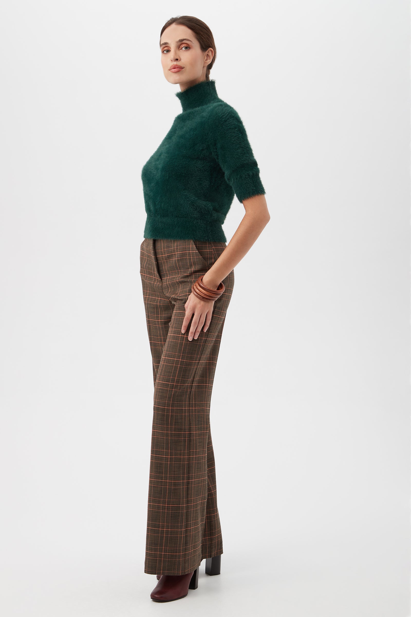 A woman in profile wears the Mali Fuzzy Sweater in dark green with brown plaid high-waisted pants, burgundy heeled boots, and brown bangles, standing against a plain white background.
