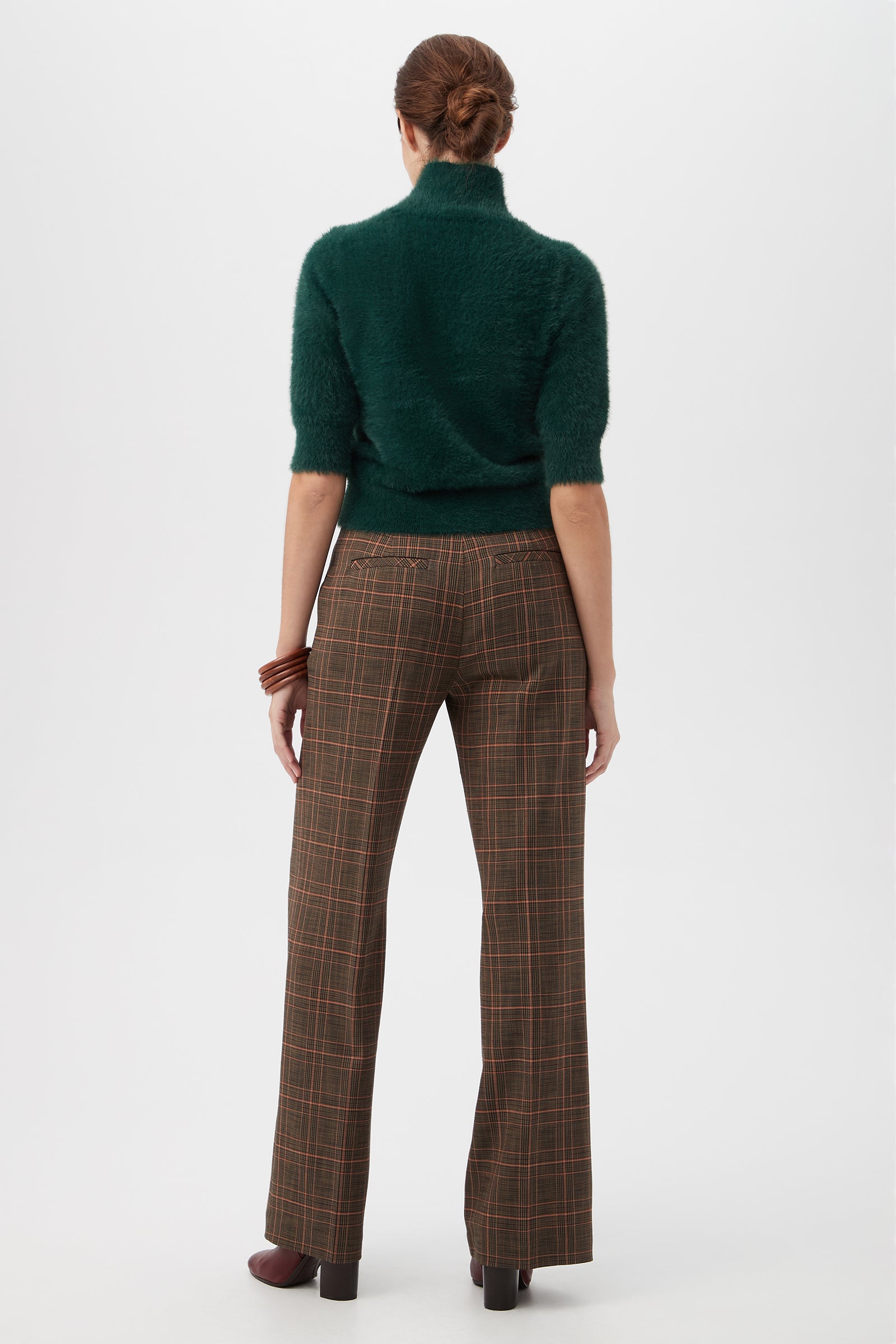 A woman with her hair in a neat bun faces away, wearing the Mali Fuzzy Sweater in green, brown plaid wide-leg pants, brown platform shoes, and a brown bracelet against a plain white background.