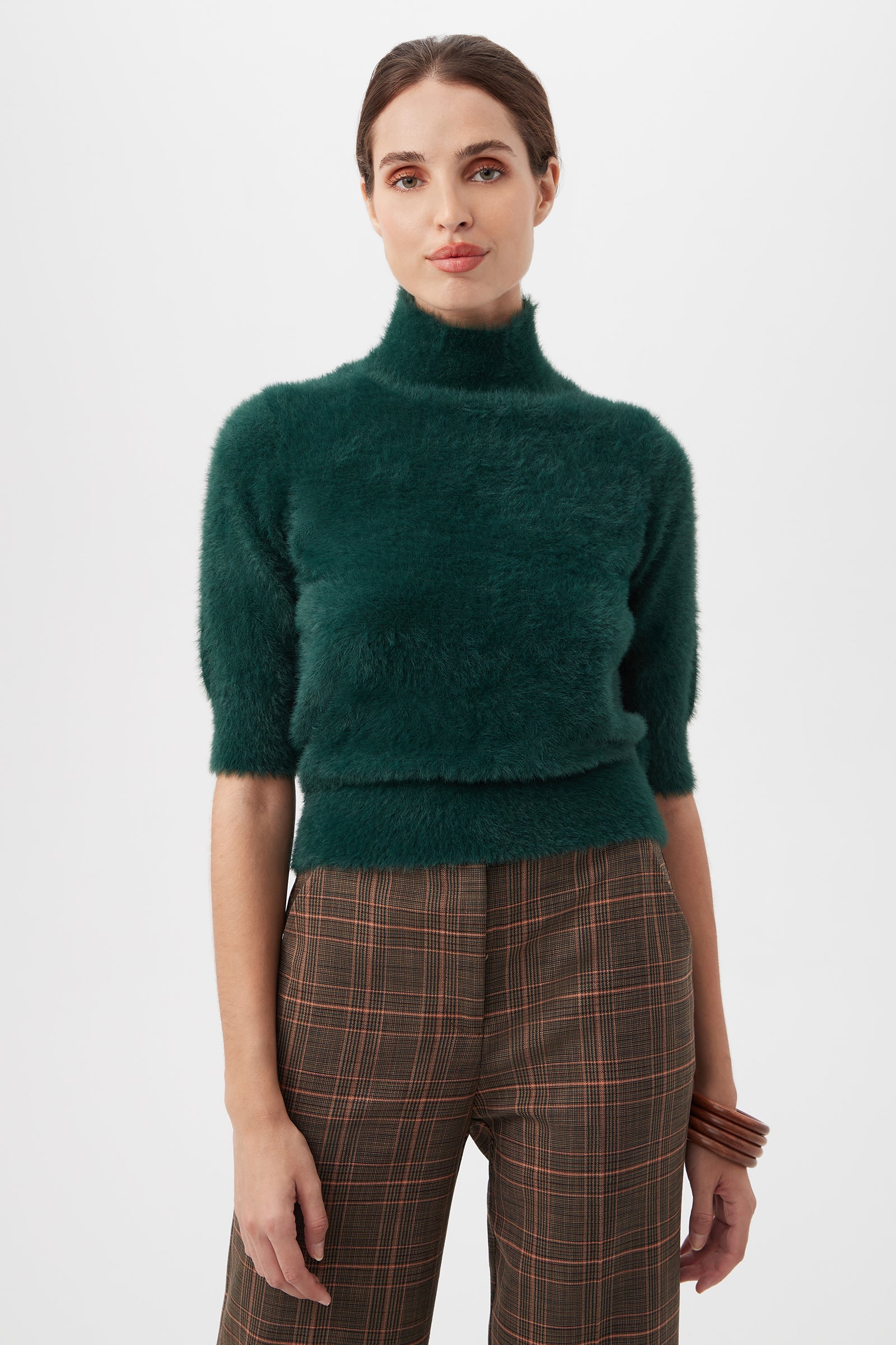 A woman wears the Mali Fuzzy Sweater in Victoria Lake, featuring a dark green turtleneck with short sleeves, paired with brown plaid trousers against a plain white background.