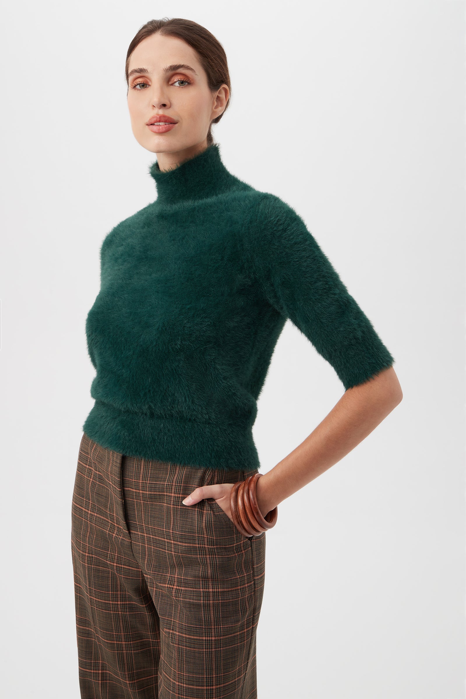 A woman wears the Mali Fuzzy Sweater, a dark green turtleneck with elbow-length sleeves, paired with brown plaid trousers, standing with one hand on her hip against a plain light background.