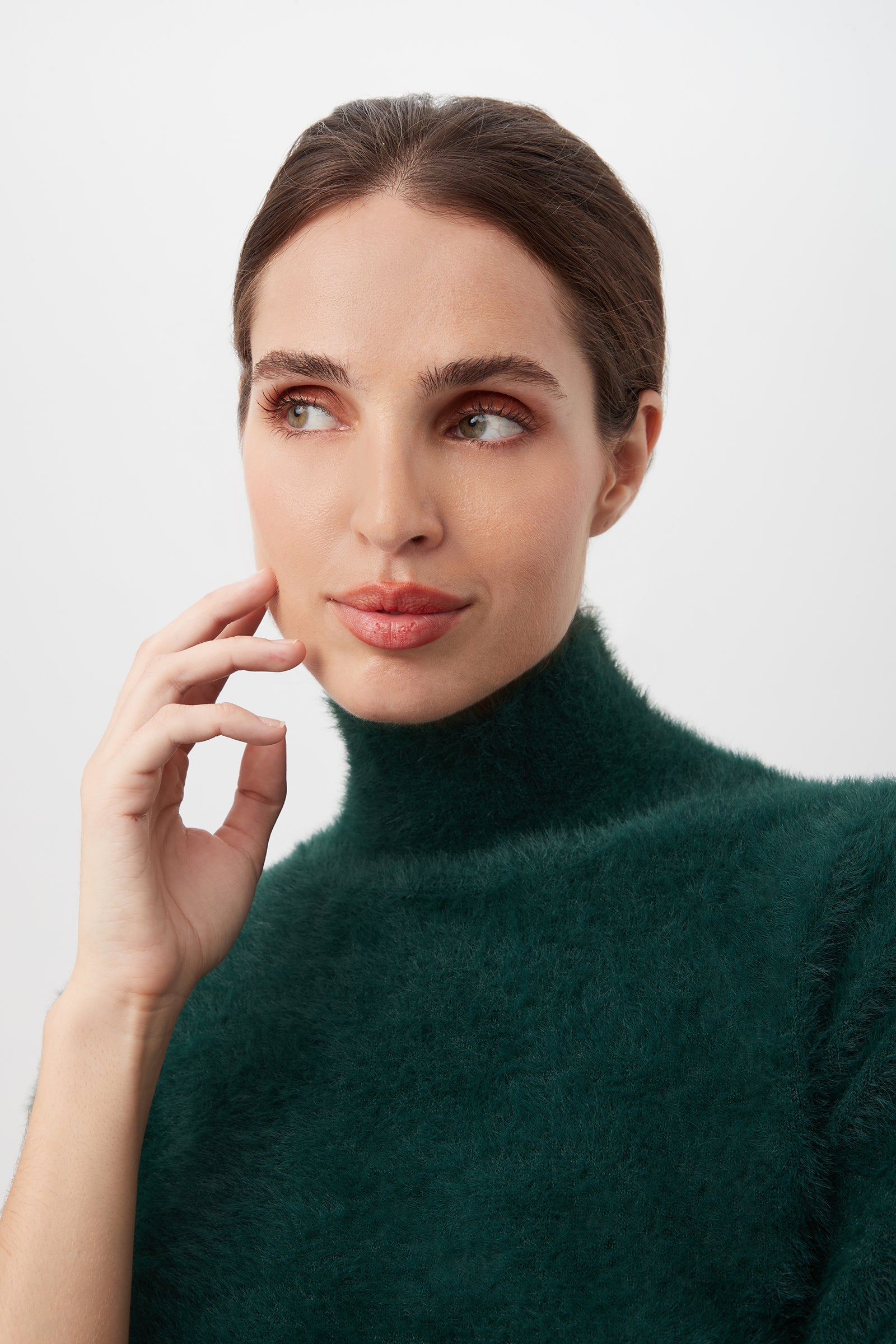 A woman with fair skin and brown hair pulled back wears the Mali Fuzzy Sweater in dark green, gazing to the side with a gentle smile and her hand touching her cheek against a white background.