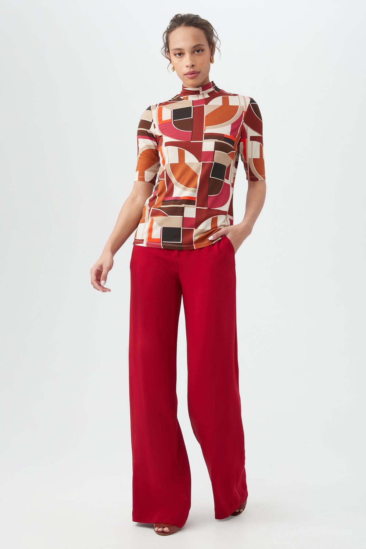 A person stands against a plain background wearing the Pierce 3 TopÃ¢â‚¬â€a geometric patterned earth-tone topÃ¢â‚¬â€with high-waisted, wide-leg red pants and one hand in their pocket.