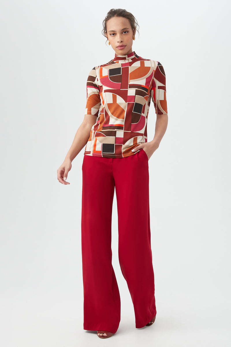 A person stands against a plain background wearing the Pierce 3 TopÃ¢â‚¬â€a geometric patterned earth-tone topÃ¢â‚¬â€with high-waisted, wide-leg red pants and one hand in their pocket.