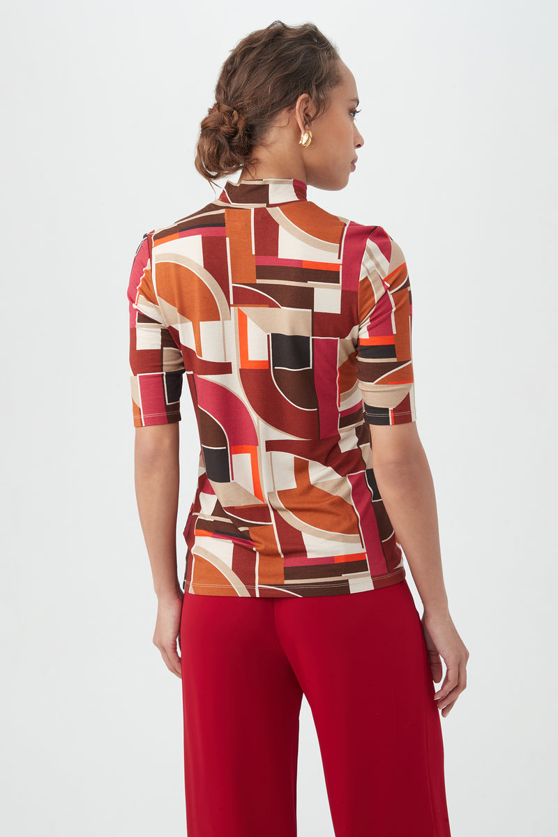 A woman with braided hair faces away, wearing the Pierce 3 TopÃ¢â‚¬â€a geometric-patterned top in brown, red, orange, and creamÃ¢â‚¬â€paired with solid red pants against a plain white background.