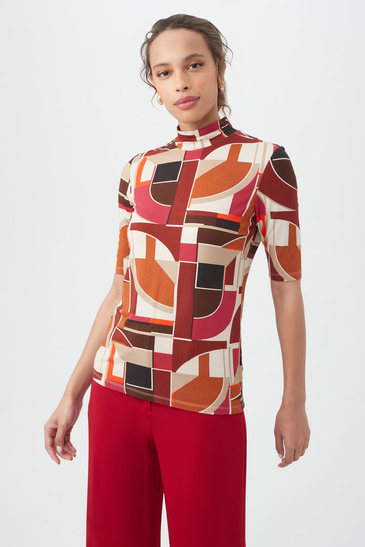 A woman stands against a plain white background wearing the Pierce 3 TopÃ¢â‚¬â€an abstract geometric print, high-neck, short-sleeved shirt in red, brown, cream, and blackÃ¢â‚¬â€paired with red pants.