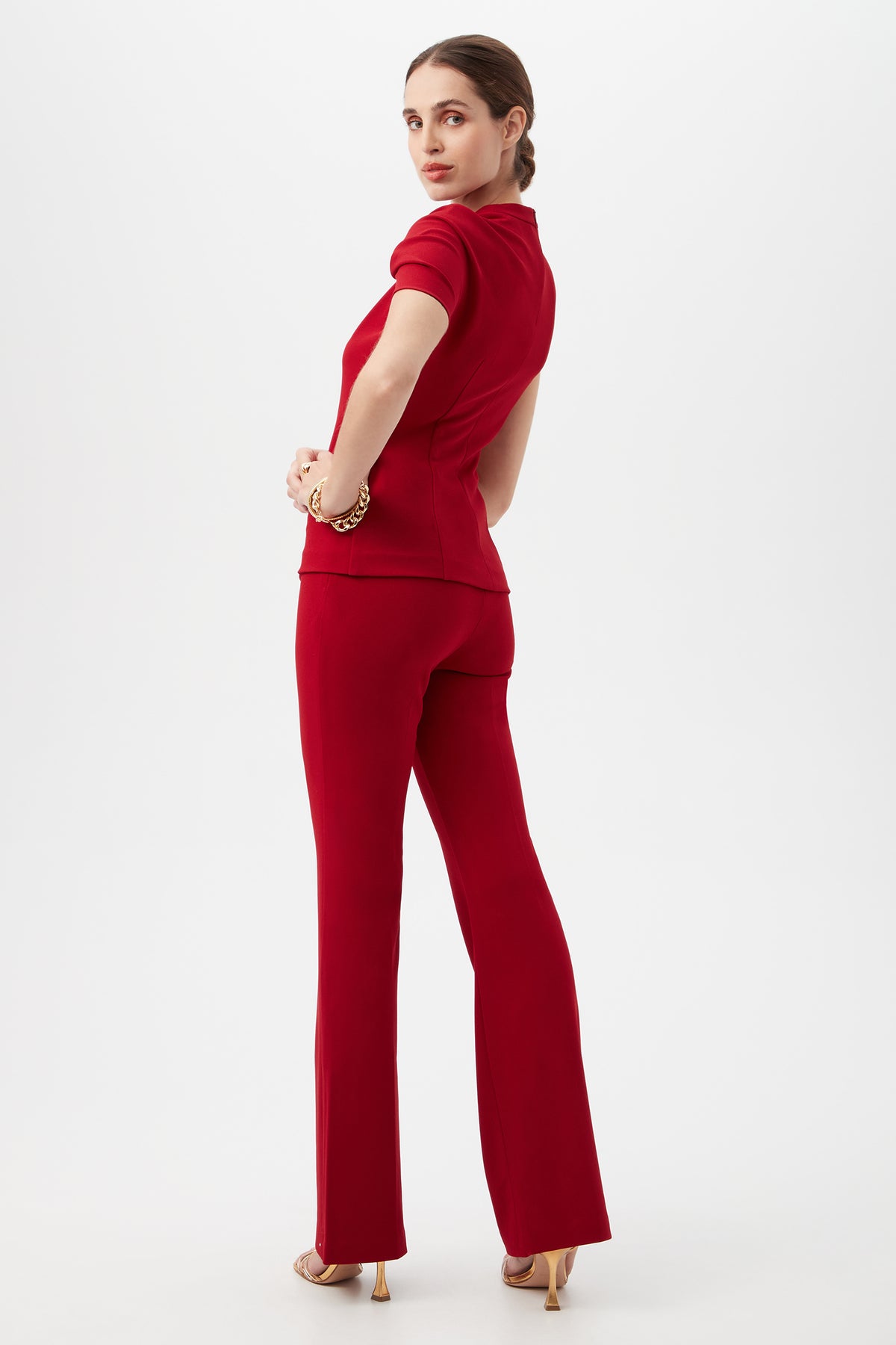 A woman stands turned to the side, looking over her shoulder, wearing the Keshi Asymmetrical Sleeve Top with Shoulder Twist Detail in red and matching flared pants, gold bracelets, and nude heels against a plain white background.