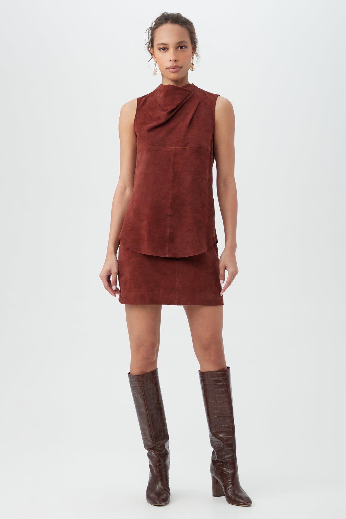 A woman stands against a white background wearing a rust-colored sleeveless suede top with an asymmetrical collar, the High Waisted Rico 2 Skirtâ€”a matching mini skirtâ€”and dark brown knee-high boots.