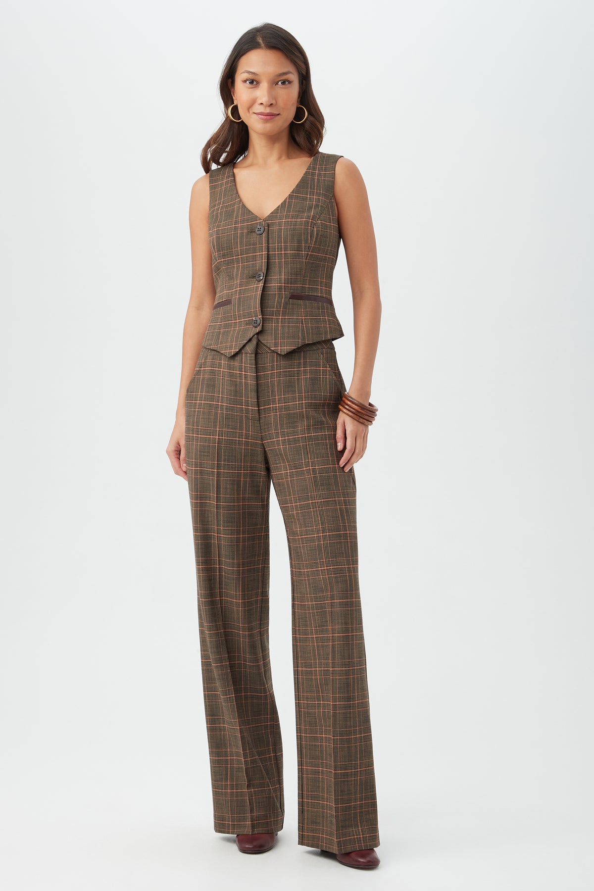 A woman with long hair, hoop earrings, and bangles stands against a white background wearing the Kofi Vest with matching brown plaid wide-leg pants and maroon shoes.