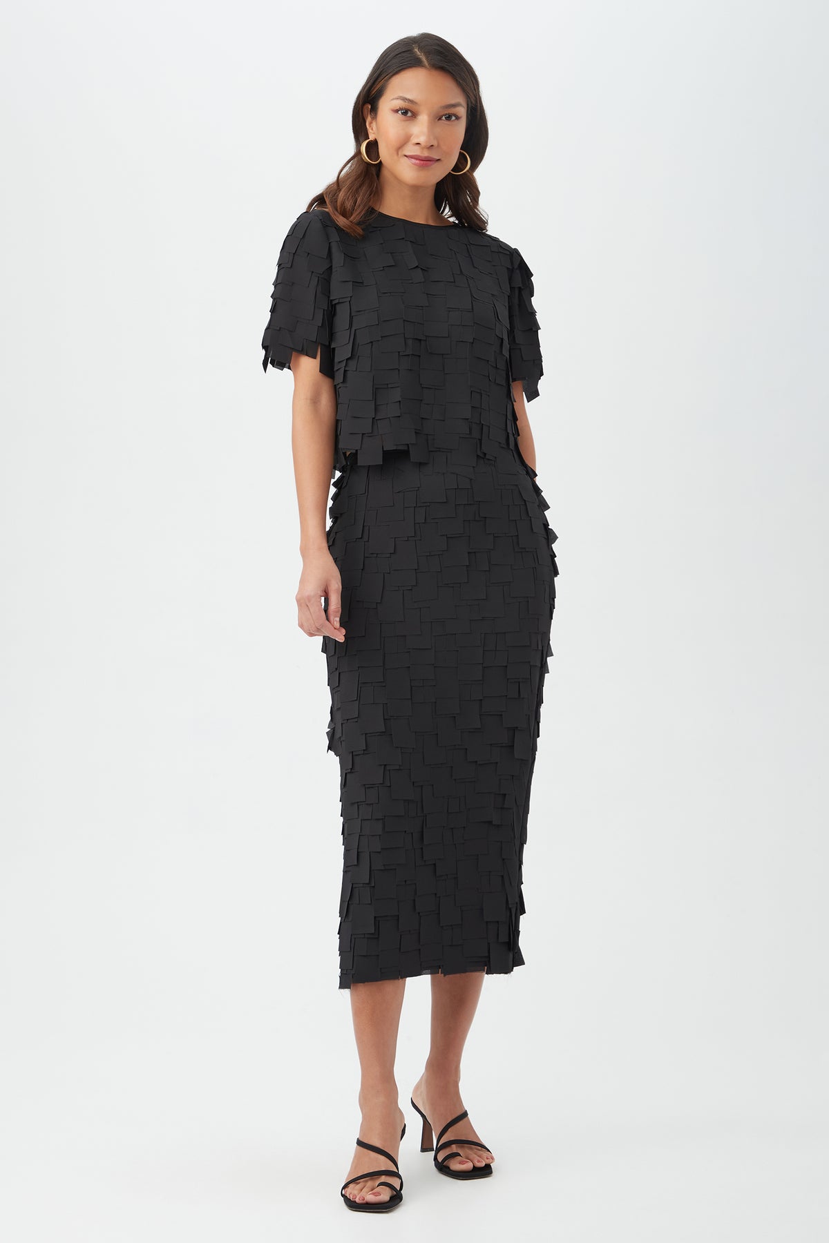 A woman stands against a plain background wearing the Serilda Short-Sleeve Top - Flutter Chiffon, paired with black strappy heels. She has wavy hair and hoop earrings.
