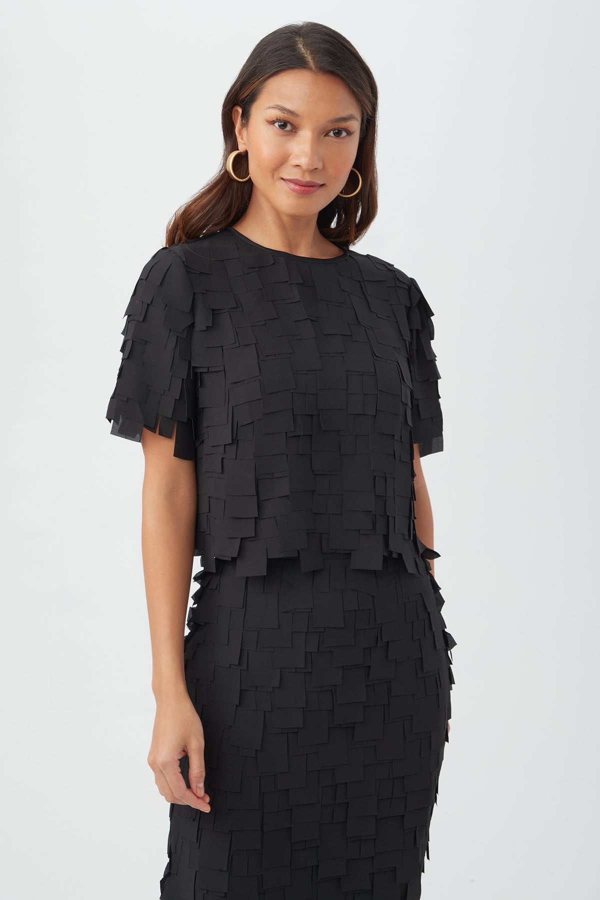 A woman in the black Serilda Short-Sleeve Top - Flutter Chiffon, styled with large gold hoop earrings and long wavy hair, stands against a plain white background, smiling slightly.