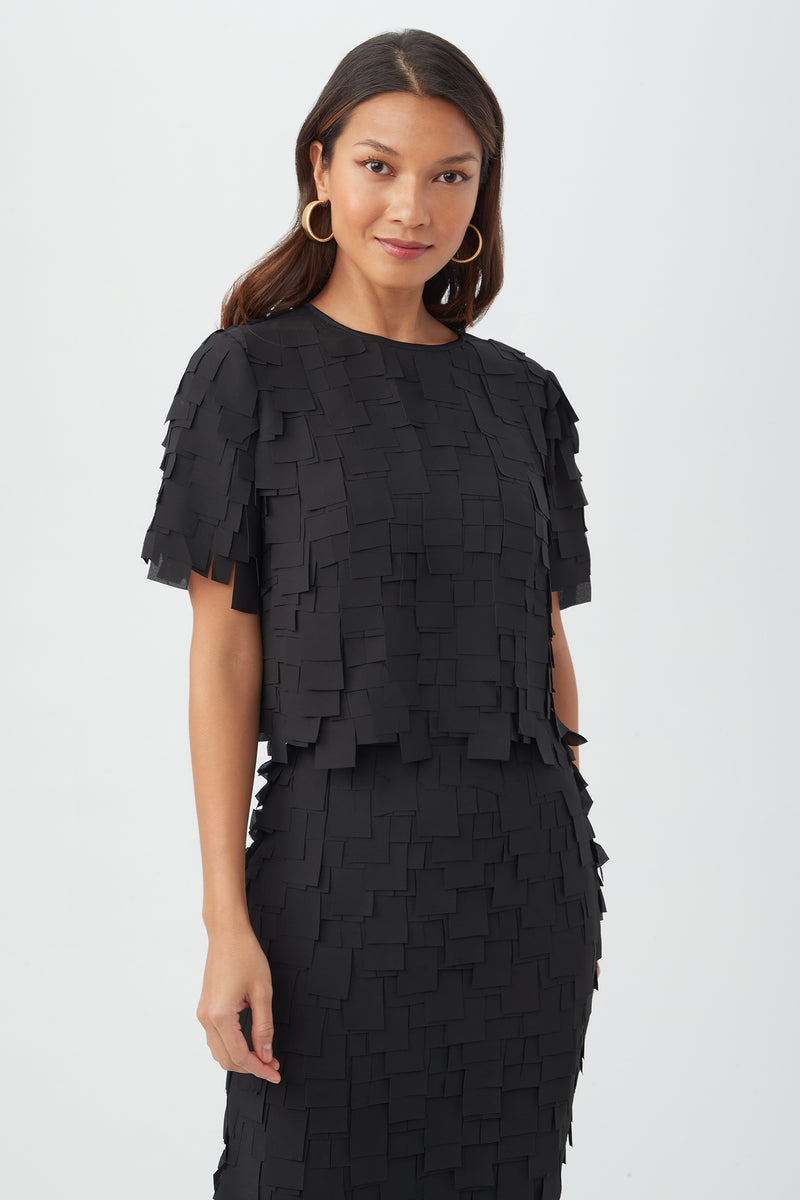 A woman in the black Serilda Short-Sleeve Top - Flutter Chiffon, styled with large gold hoop earrings and long wavy hair, stands against a plain white background, smiling slightly.