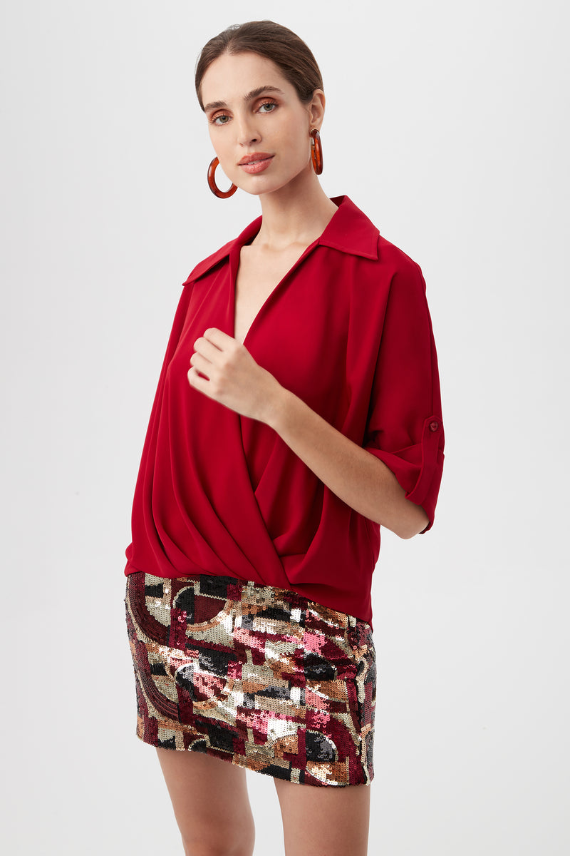 A woman wears the Otto Top, a deep red, long-sleeved blouse with a draped front, paired with a shimmering multicolored sequin mini skirt. Her hair is pulled back, she wears hoop earrings, and stands against a plain light background.