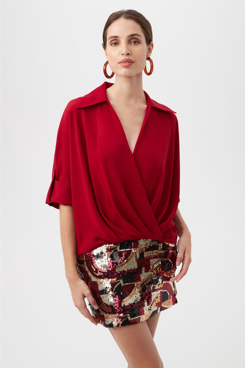 A woman poses against a white background wearing the Otto Top in Roselle Red with long sleeves and rolled cuffs, paired with a shiny sequin mini skirt. Her hair is pulled back and she accessorizes with large red hoop earrings.