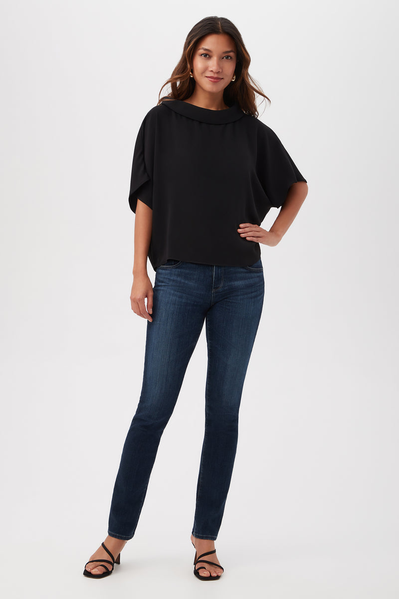 A woman with long brown hair stands against a white background, wearing the Honeysuckle Boatneck Dolman-Sleeve Top in black, paired with dark blue skinny jeans and black strappy heels. She poses confidently with one hand on her hip.