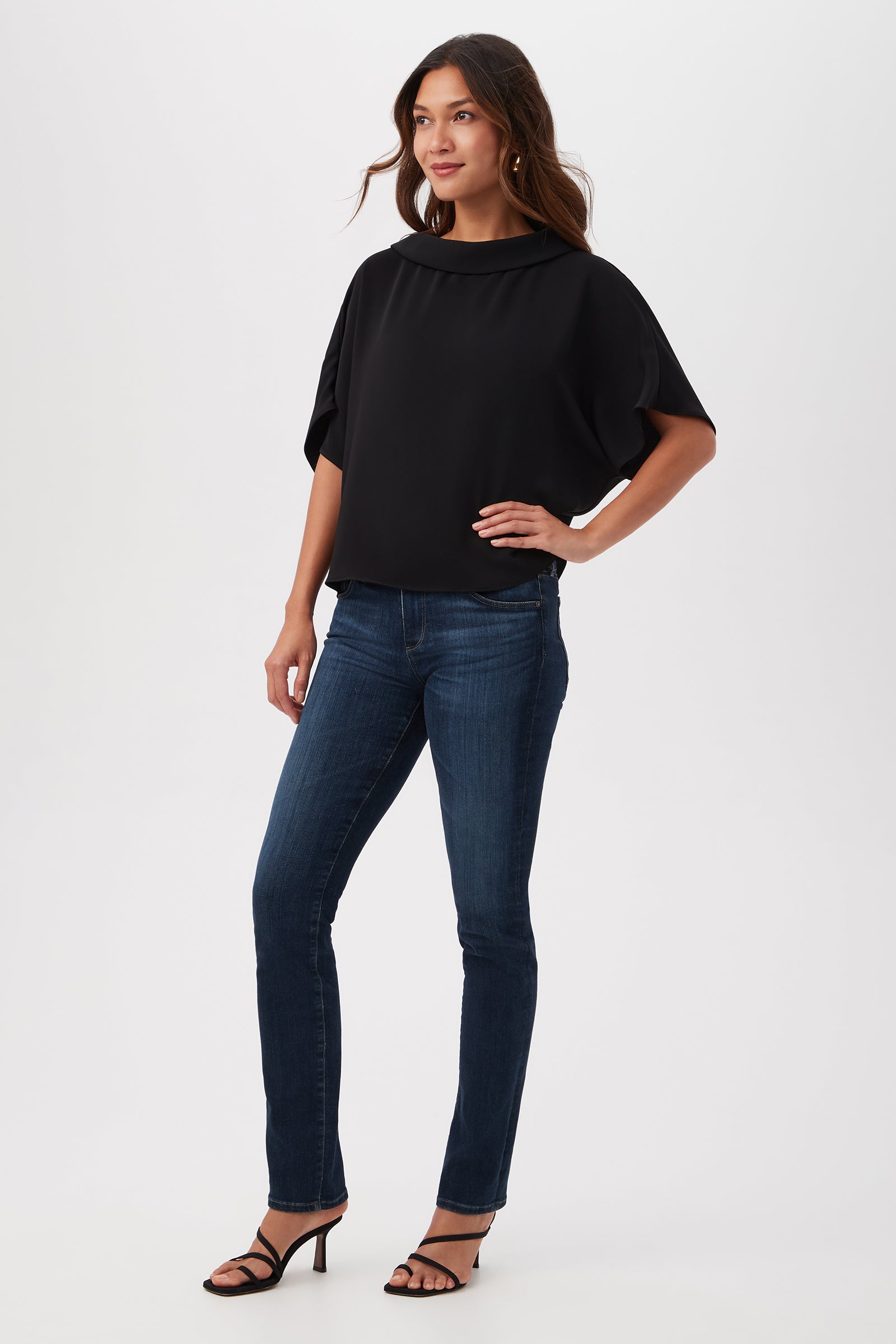 A woman with long, wavy hair stands against a white background, smiling slightly with one hand on her hip. She wears the Honeysuckle Boatneck Dolman-Sleeve Top in black, paired with dark blue jeans and black heeled sandals.