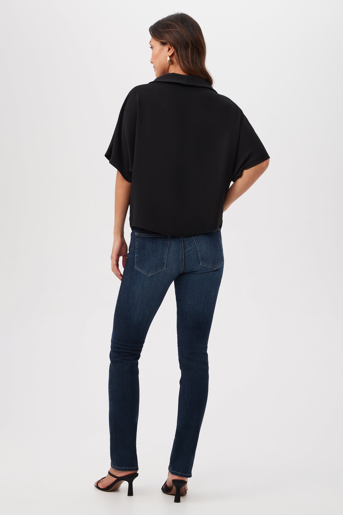 A woman stands facing away, wearing the Honeysuckle Boatneck Dolman-Sleeve Top in black, fitted blue jeans, and black heeled sandals against a plain white background.