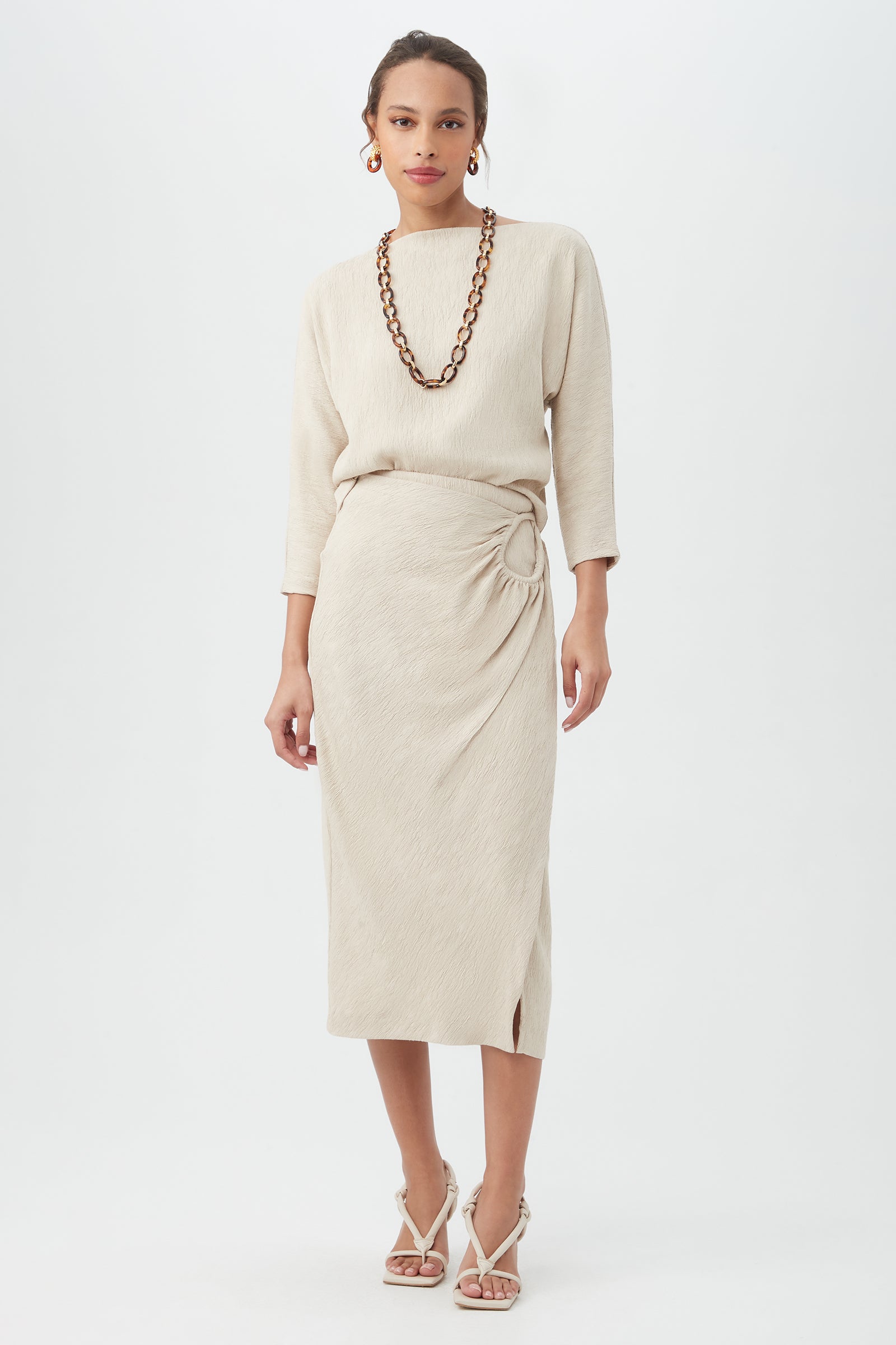 A woman poses against a white background wearing the cream Imma Top with matching long-sleeve midi skirt featuring a knotted detail, styled with strappy heels, a chunky necklace, and hoop earrings.