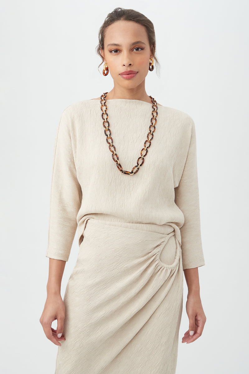 A woman wears the Imma Top in Sahara Stone with a matching skirt featuring a side knot, accessorized with a chunky tortoiseshell necklace and hoop earrings, standing against a plain light background.
