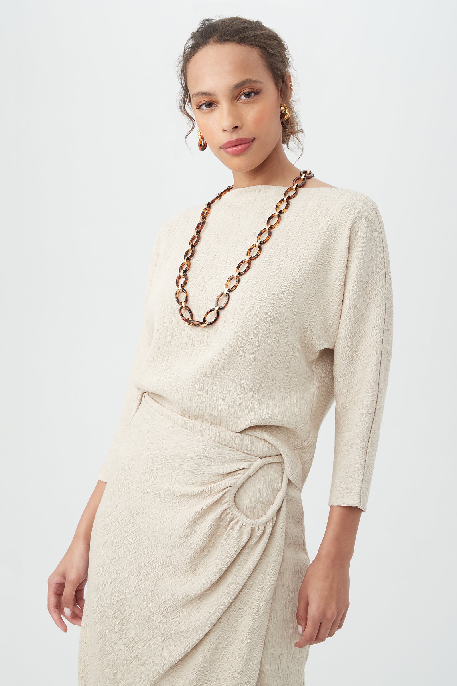 A woman wears the Imma Top—a beige, textured long-sleeve top paired with a matching skirt featuring gathered detail and a circular accent—styled with a chunky brown tortoiseshell necklace and gold hoop earrings.