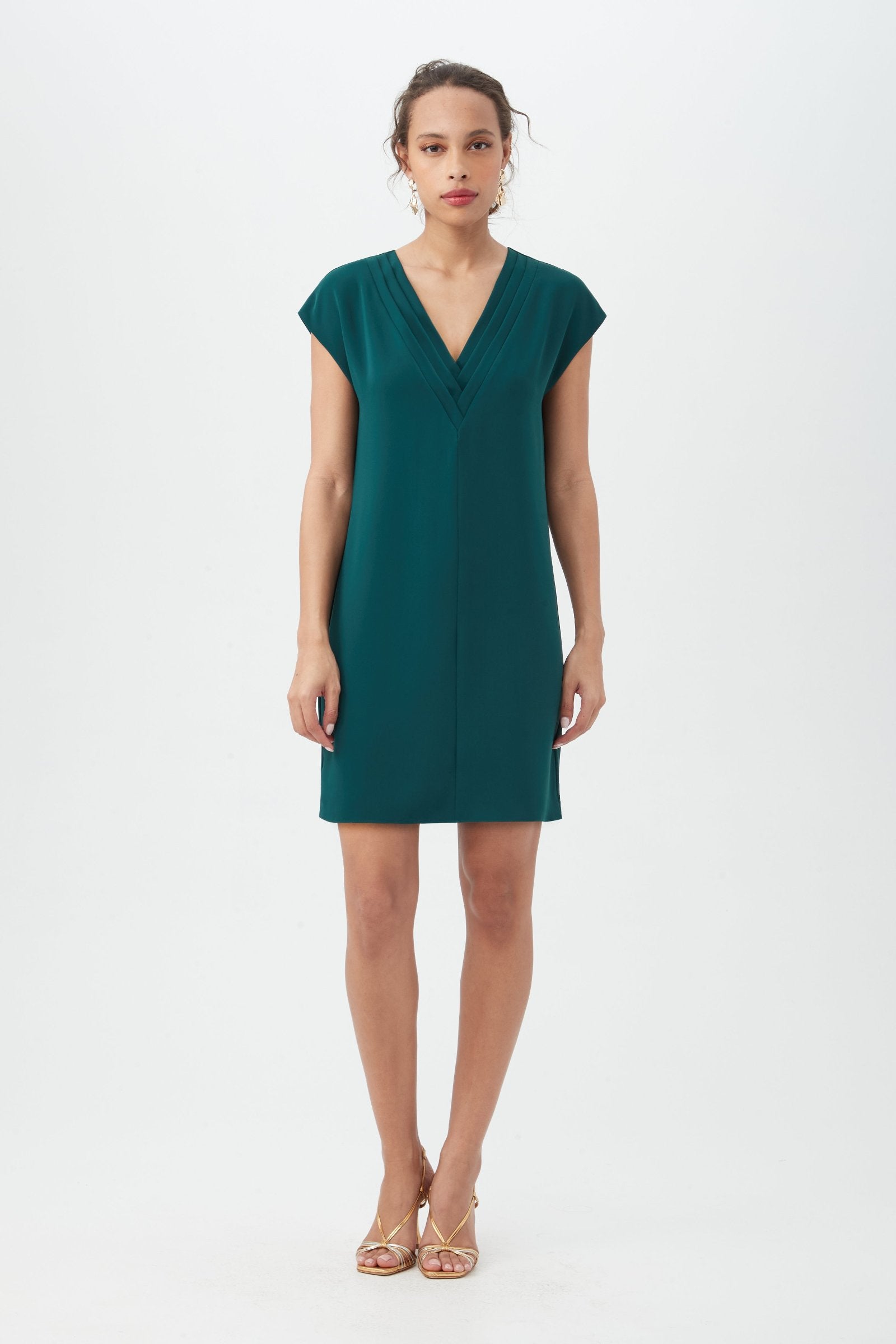 A woman stands against a white background wearing the Zula Dress in Victoria Lake, a short, dark teal V-neck style with cap sleeves, paired with gold strappy heels. Her curly hair is pulled back as she faces forward with a neutral expression.