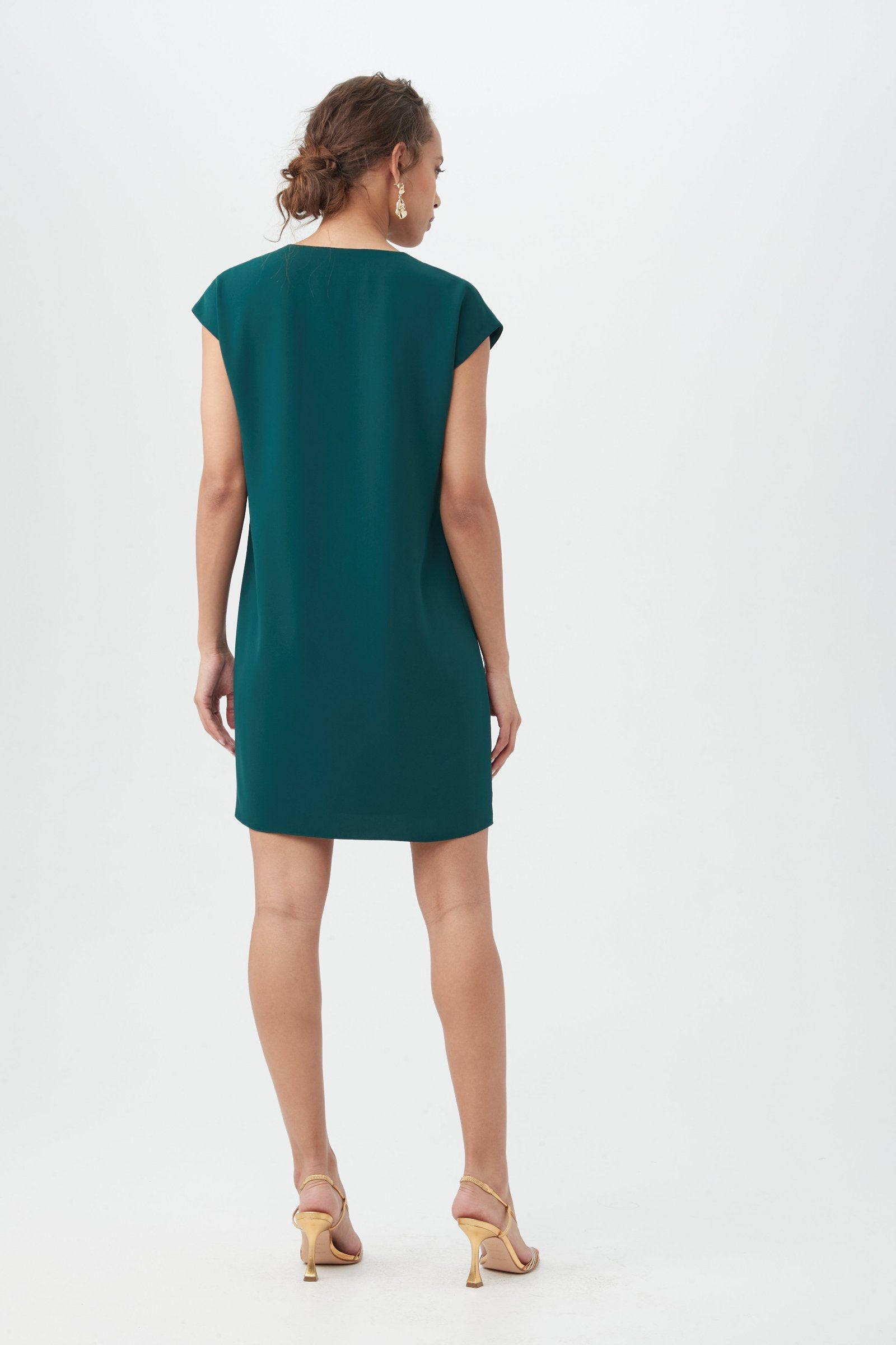 A woman faces away, wearing the Zula Dressâ€”a short-sleeved, knee-length teal dressâ€”with beige high-heeled sandals against a plain white background.