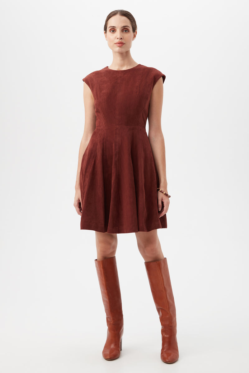 A woman poses against a white background, wearing the sleeveless, knee-length Sabi Dress in mahogany with a fitted waist and flared skirt, paired with tall brown leather boots.