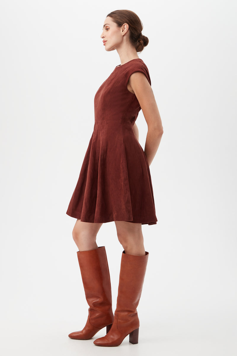 A woman stands in profile against a plain white background, wearing the Sabi DressÃ¢â‚¬â€short-sleeved, knee-length, and burgundyÃ¢â‚¬â€with her hair in a neat bun and tall brown heeled boots.