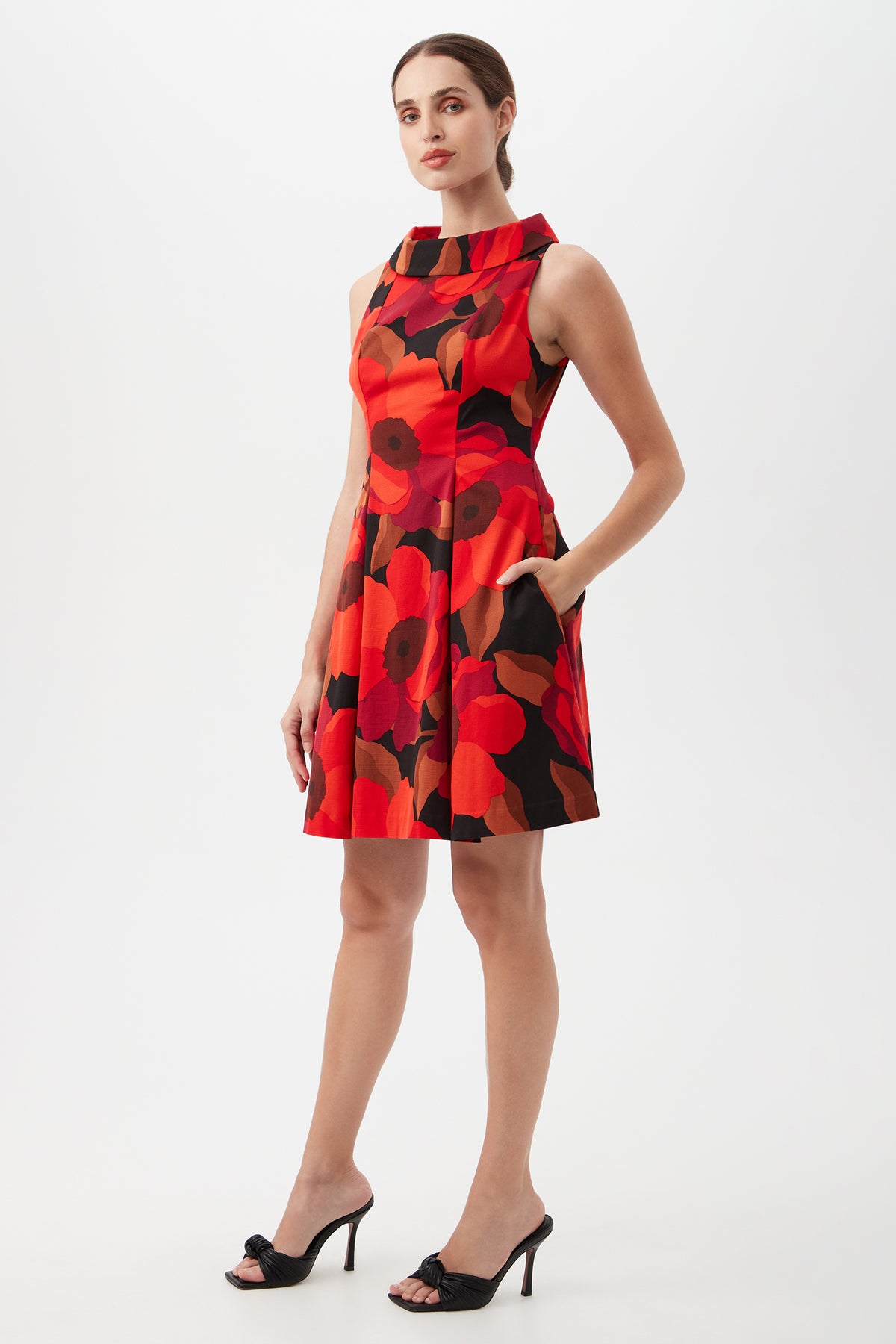 A woman models the Sabar Red Floral Sleeveless Cocktail Dress, a knee-length design with bold red florals, hands in pockets, paired with black open-toe heels, against a white background.
