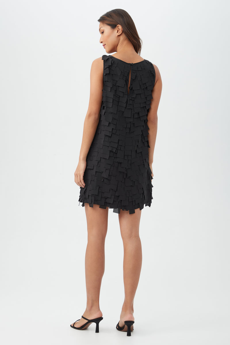 A woman faces away, wearing the Valdis Dressâ€”a black sleeveless dress with a textured, layered square patternâ€”and black heeled sandals against a plain white background.