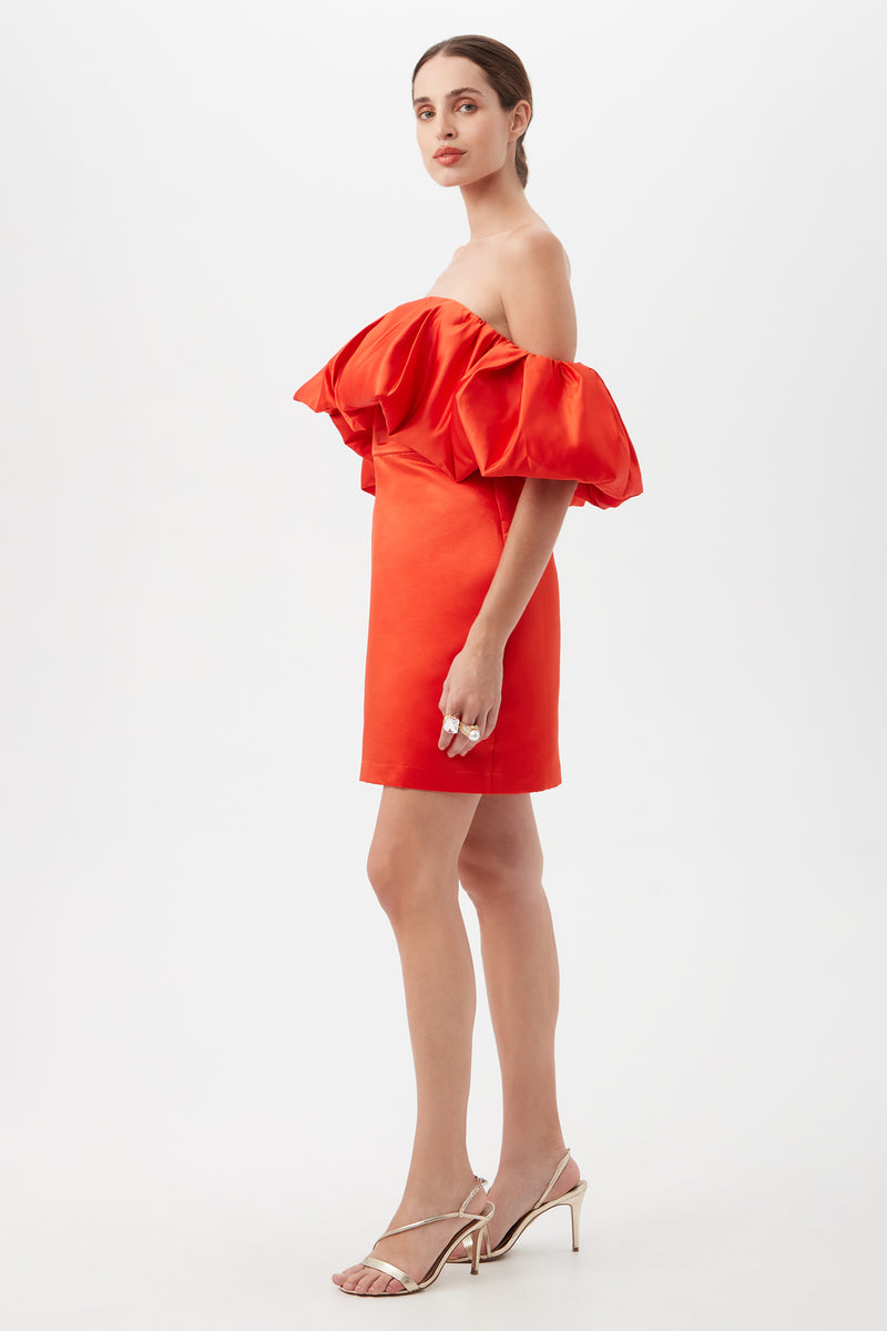 A woman stands in profile against a white background, wearing the Nia Satin Off The Shoulder Short Cocktail Dress in bright orange with puffed sleeves and heeled sandals.